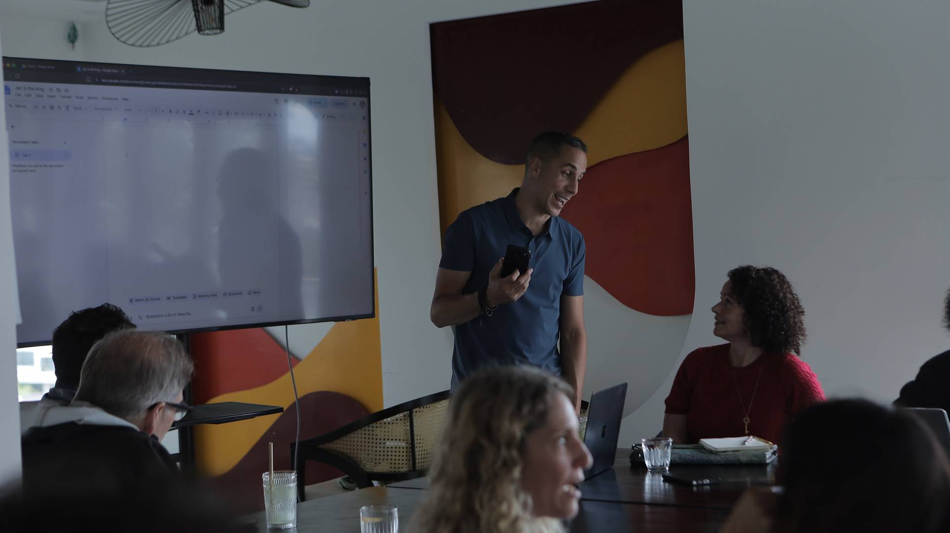 A man stands presenting to a small group in a modern office setting with colorful artwork and a large screen displaying Google Docs.