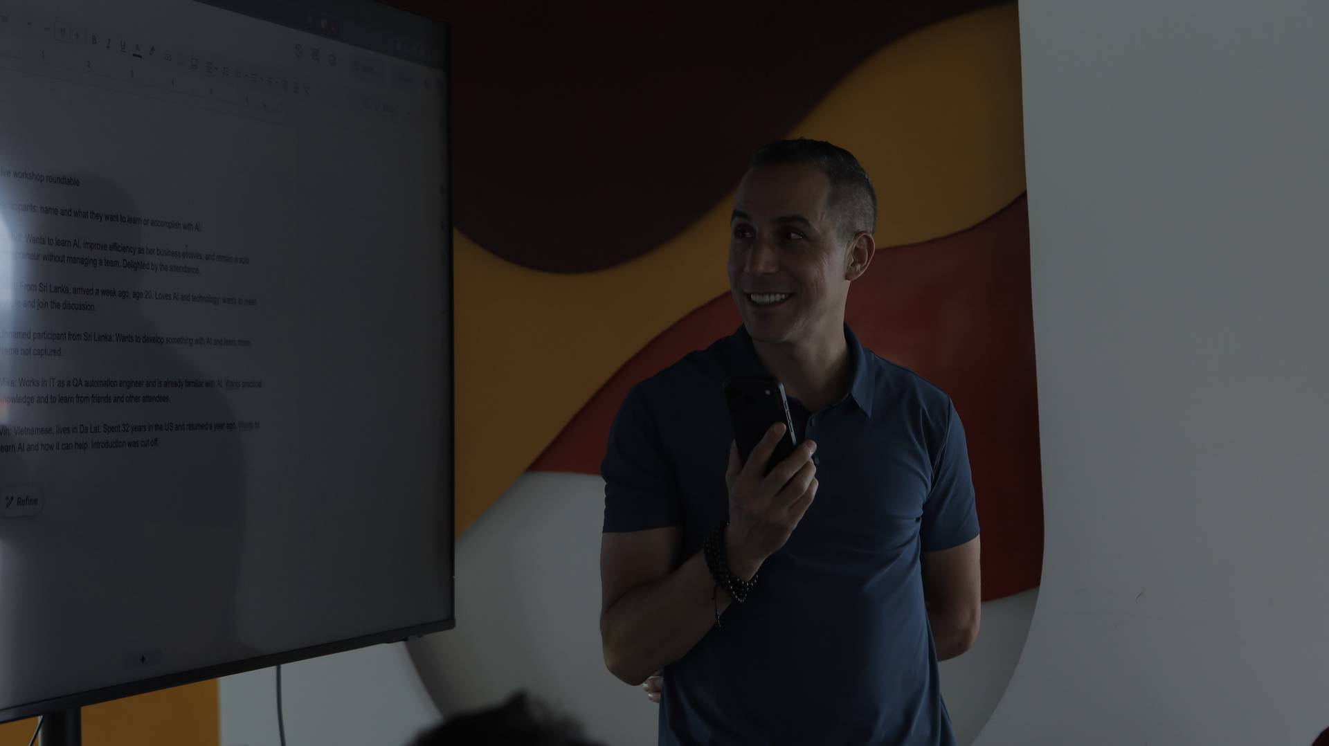 A man smiles while holding a smartphone in front of a presentation screen during a workshop roundtable discussion about AI in Đà Lạt, Vietnam.