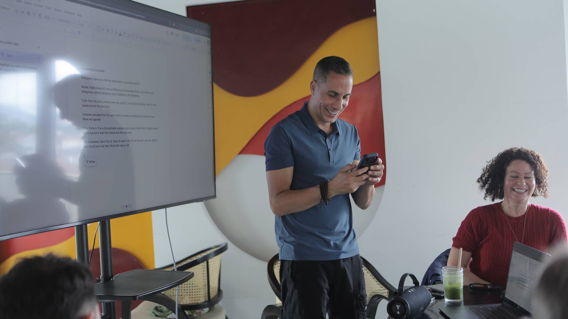 A man stands in front of a large screen displaying workshop notes during a lively roundtable discussion in Đà Lạt, Vietnam.