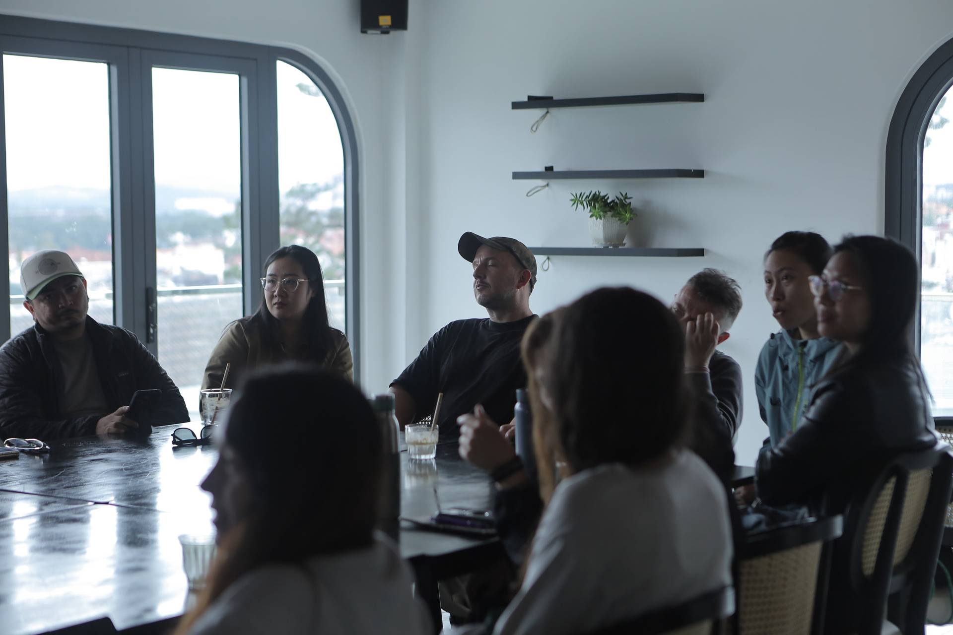 A group of people engaged in a discussion around a table with drinks, set against large windows overlooking Đà Lạt's scenic landscape.