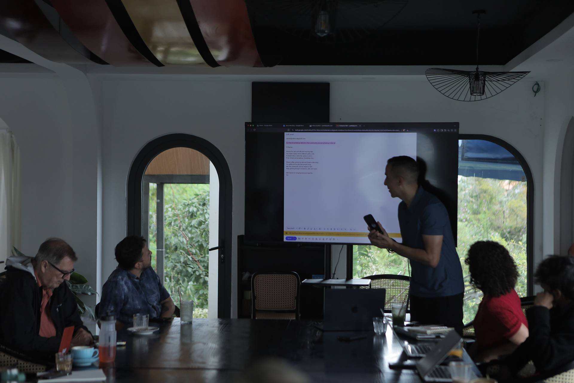 A group of people is gathered in a modern conference room in Đà Lạt, Vietnam, engaging in a presentation with a large screen displaying a document. The setting features arched windows and lush greenery outside, creating a serene atmosphere.