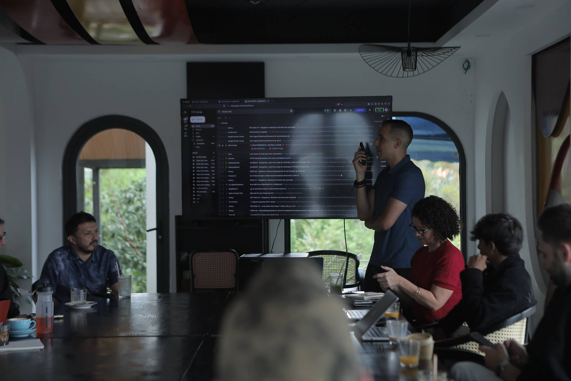 A professional meeting in a modern office space with a large screen displaying email content, surrounded by attendees engaged in discussion and taking notes.