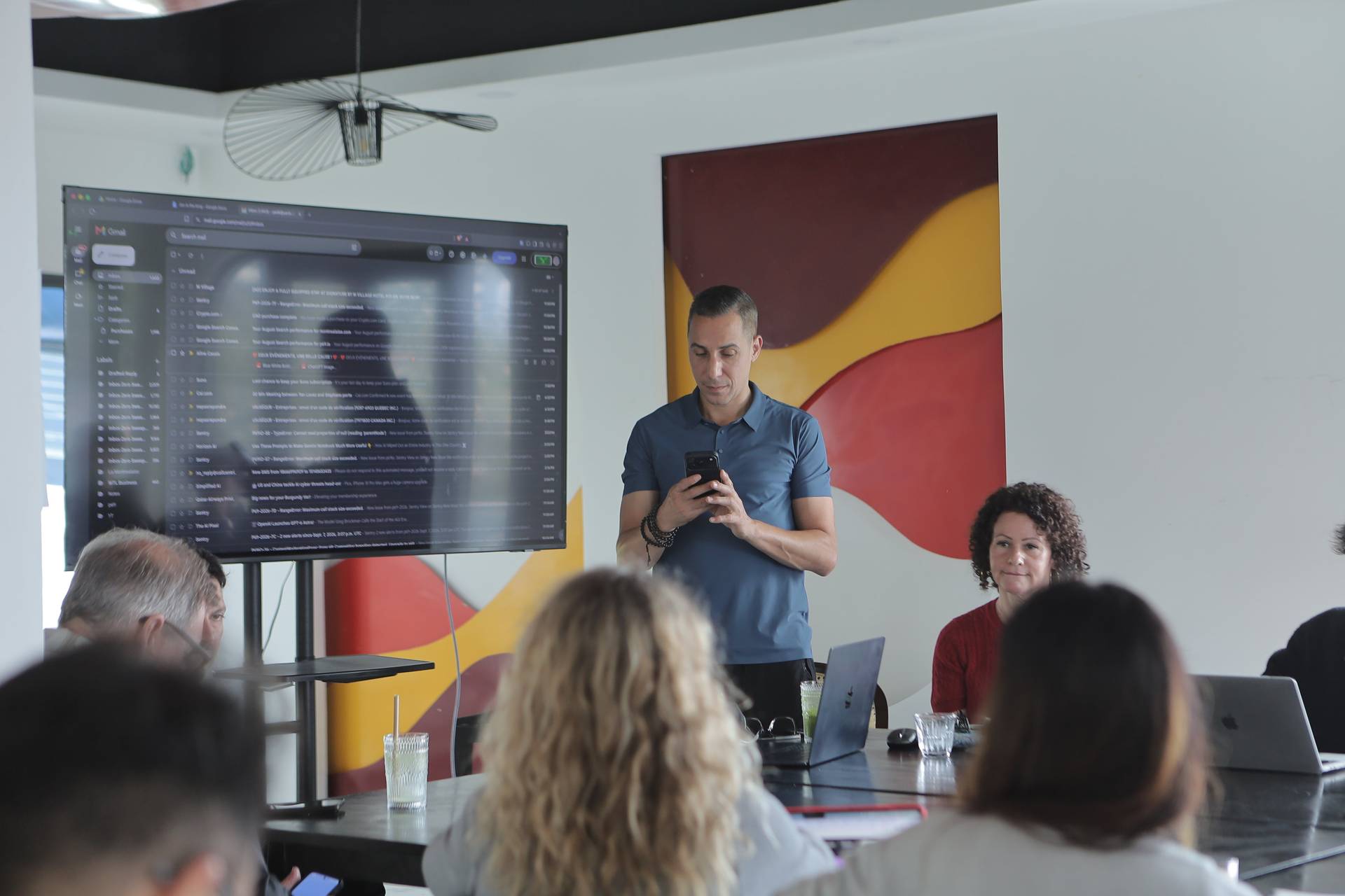 A professional meeting in Đà Lạt, Vietnam, with a presenter using a smartphone while attendees focus on a large screen displaying an email inbox.
