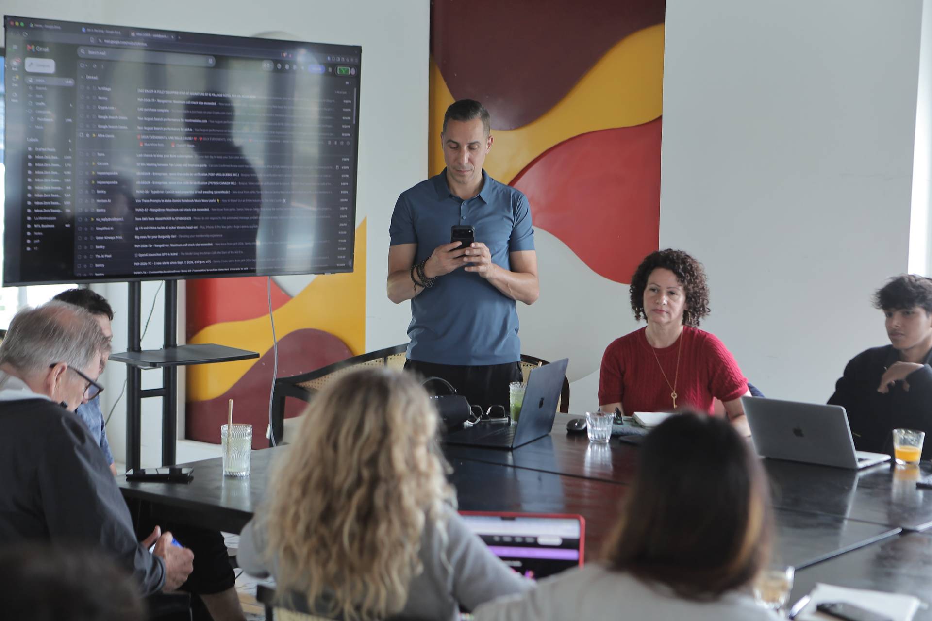 A professional meeting in a modern office setting with participants engaged in discussion around a large table equipped with laptops and refreshments.