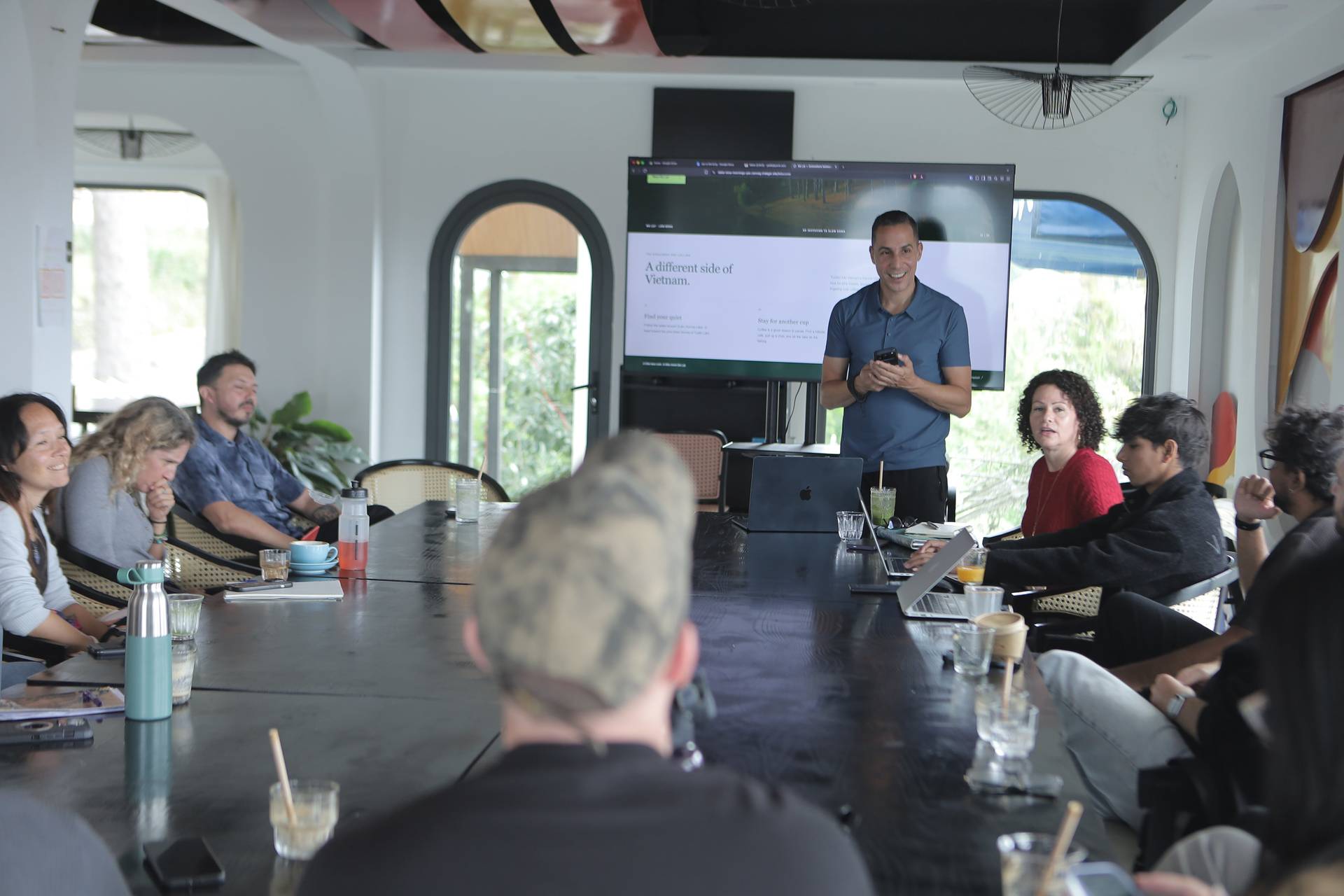 A group of people is gathered around a large table in a modern conference room in Đà Lạt, Vietnam, engaging in a presentation about 'A different side of Vietnam'. The setting includes contemporary decor and natural light streaming through large windows.