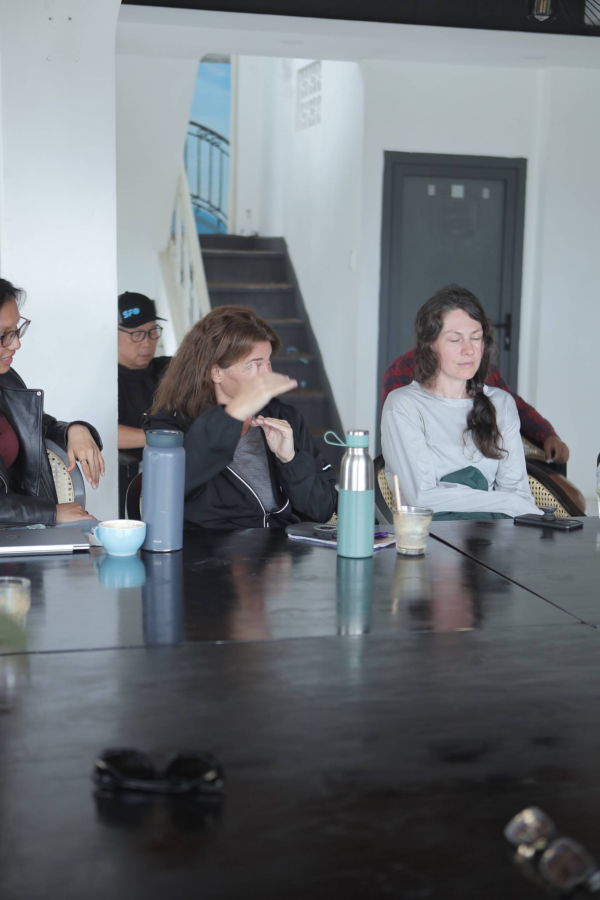 A group of people is gathered around a table in a modern indoor setting, engaging in what appears to be a casual meeting or discussion over drinks and snacks.