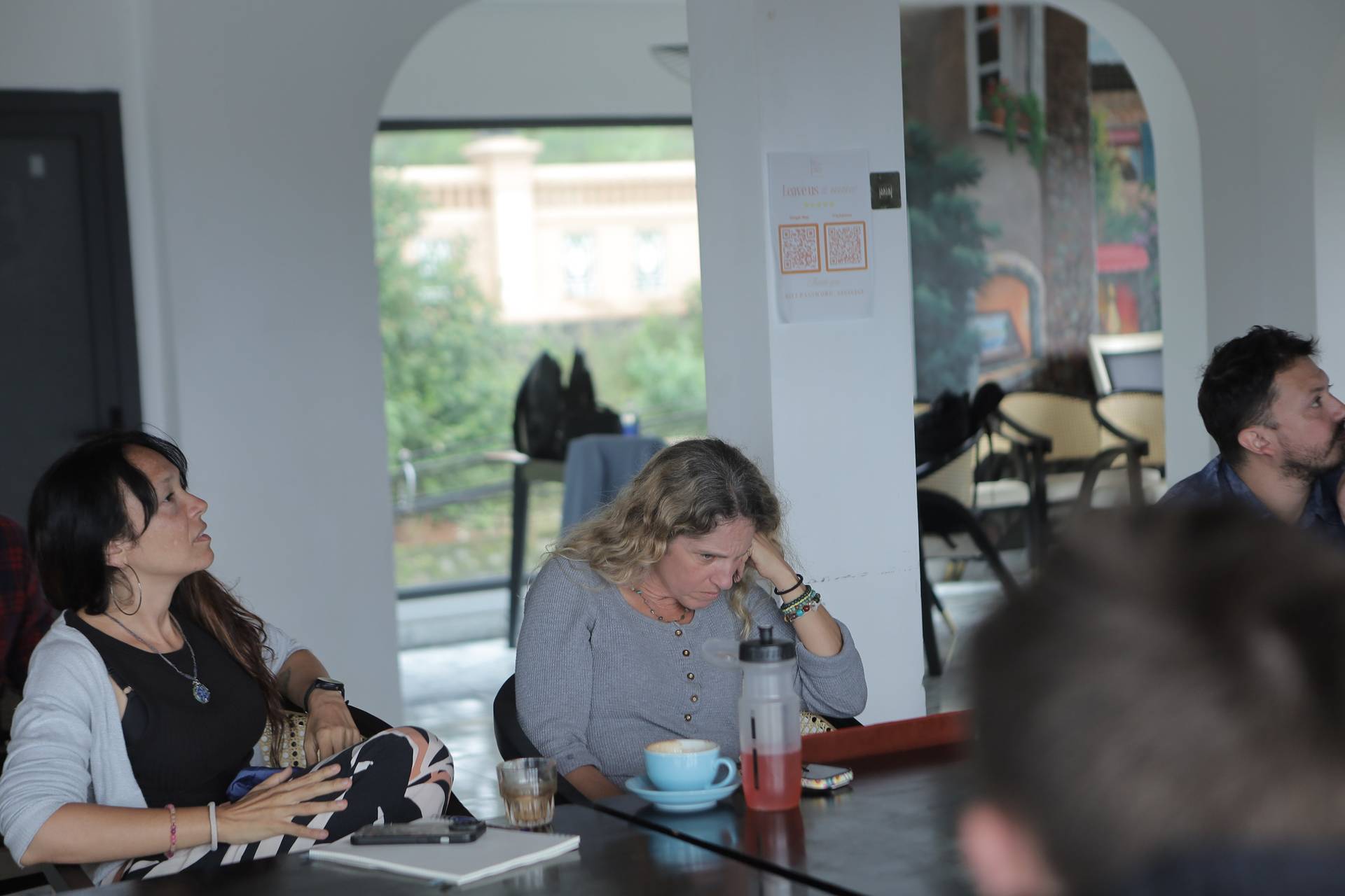 A group of people engaged in a discussion over coffee in a cozy café setting with scenic views of Đà Lạt's architecture and greenery.