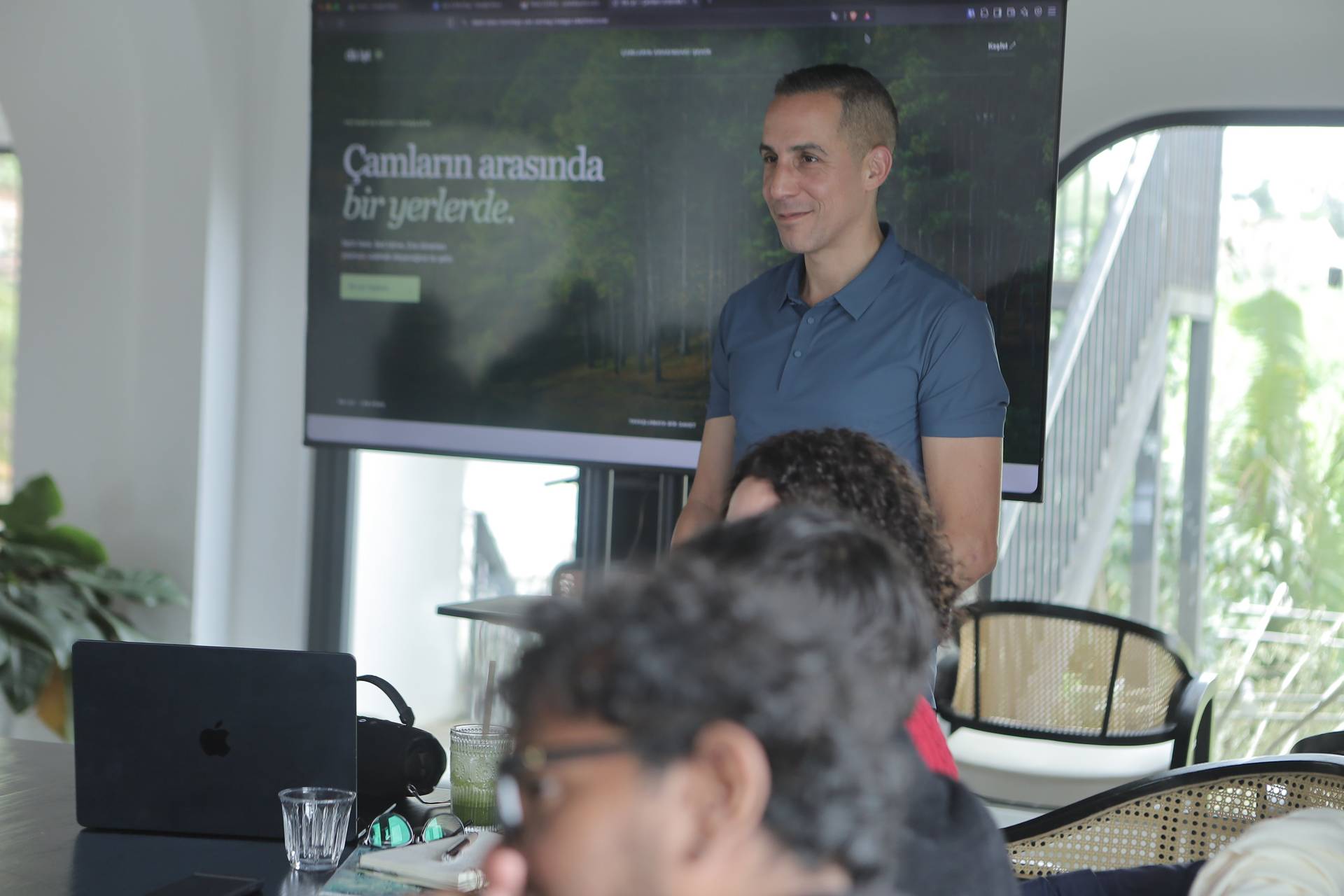 A professional presentation in a modern office setting with attendees focused on the speaker and a large screen displaying a forest-themed slide.