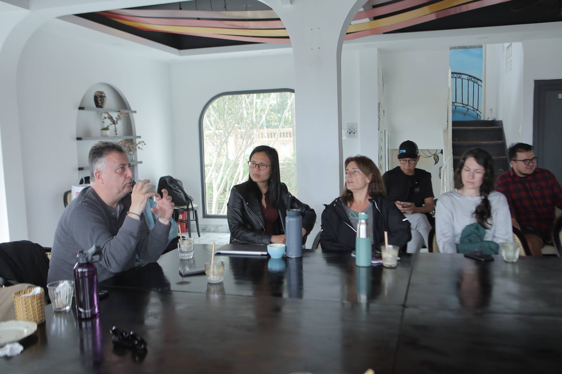 A group of people engaged in a discussion around a table in a modern café setting with large windows overlooking greenery.