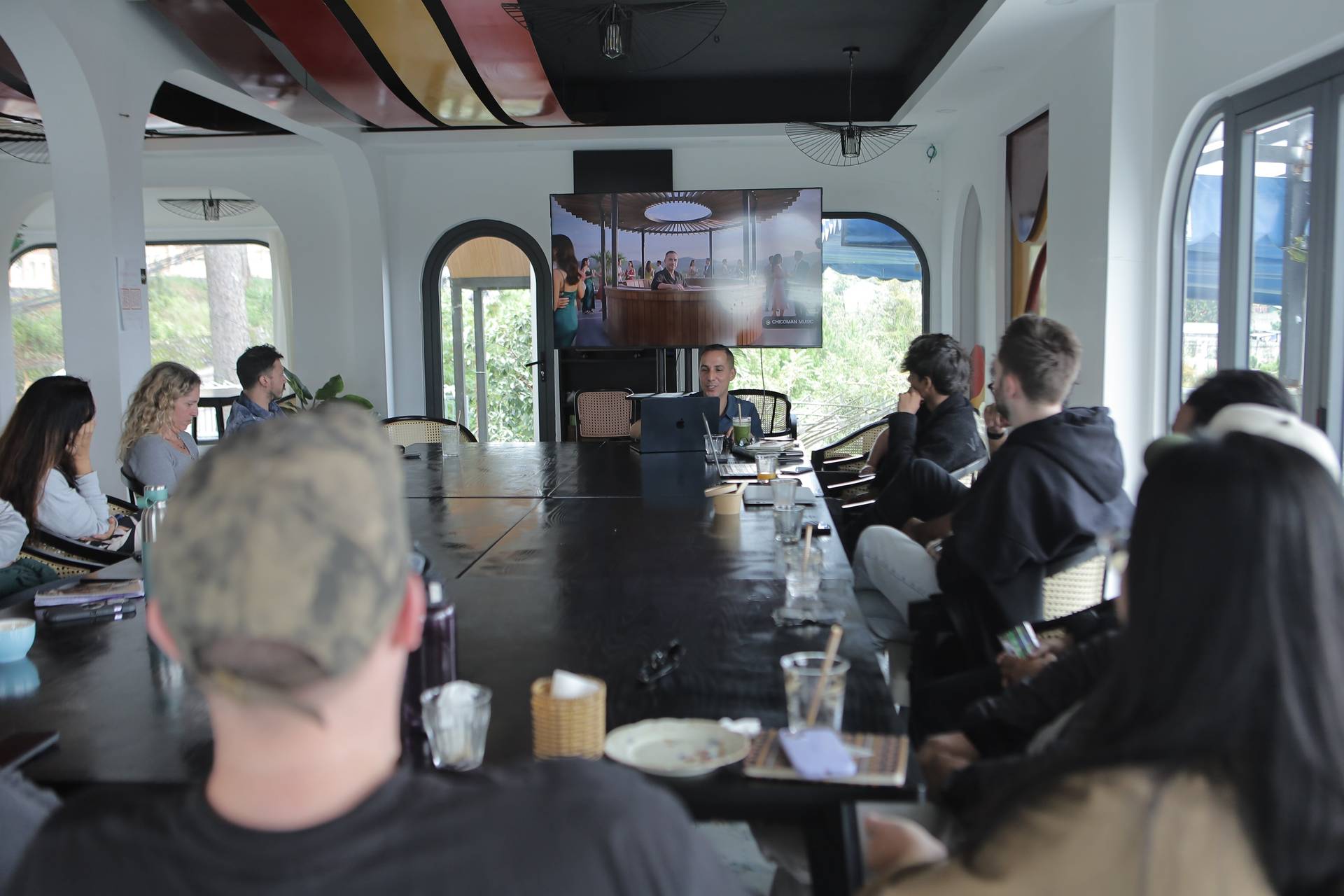 A group of people gathered around a long table in a modern Vietnamese setting, engaging in what appears to be a business or creative meeting with a scenic view outside.