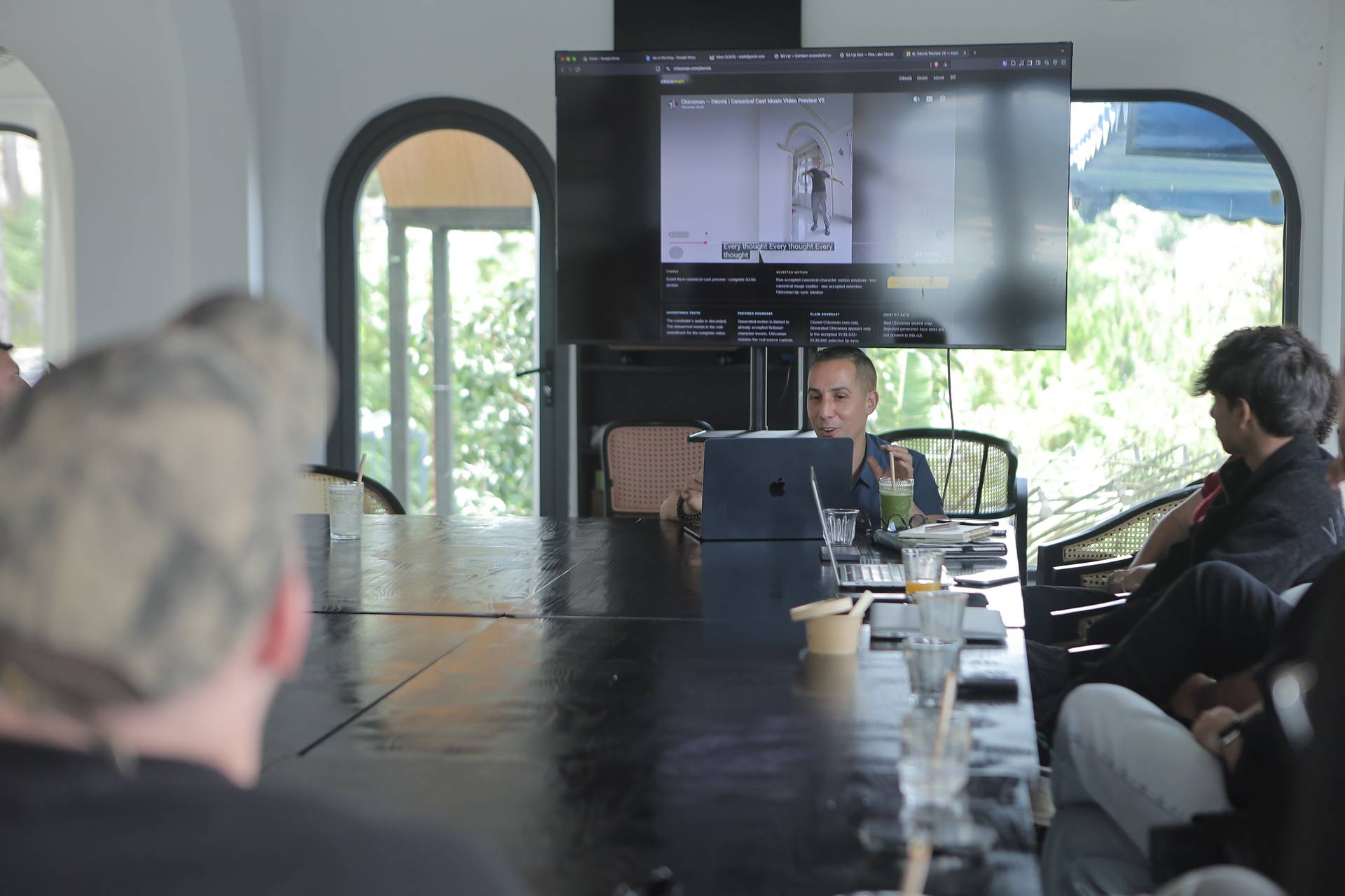 A group of people engaged in a discussion around a large table with a laptop and drinks, set against a backdrop of arched windows overlooking lush greenery.