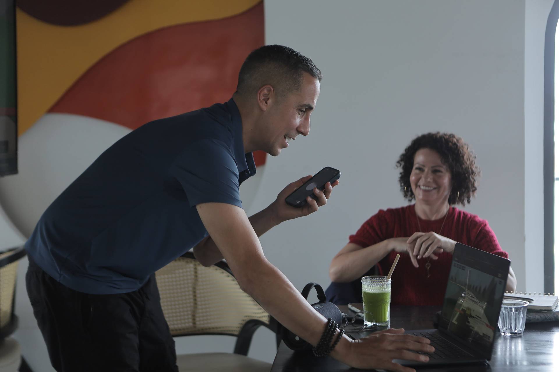 A man leans over a table in Đà Lạt, Vietnam, engaging with his phone while a woman smiles and sits at the same table, enjoying a green smoothie.