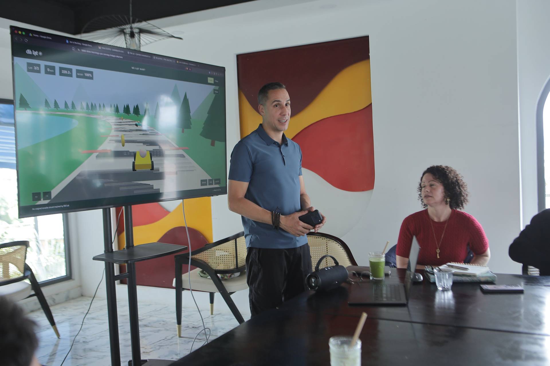 A man presents a digital racing game to an audience in a modern conference room with vibrant decor and Vietnamese branding.