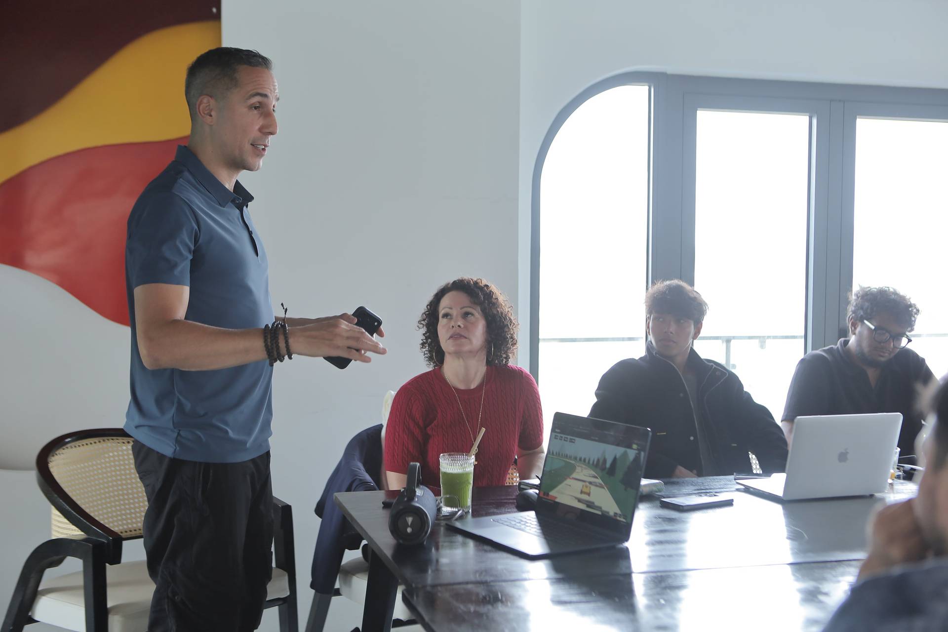 A man stands presenting to a small group seated around a table in a modern office space with large windows and colorful wall art, suggesting an engaging business or creative meeting.