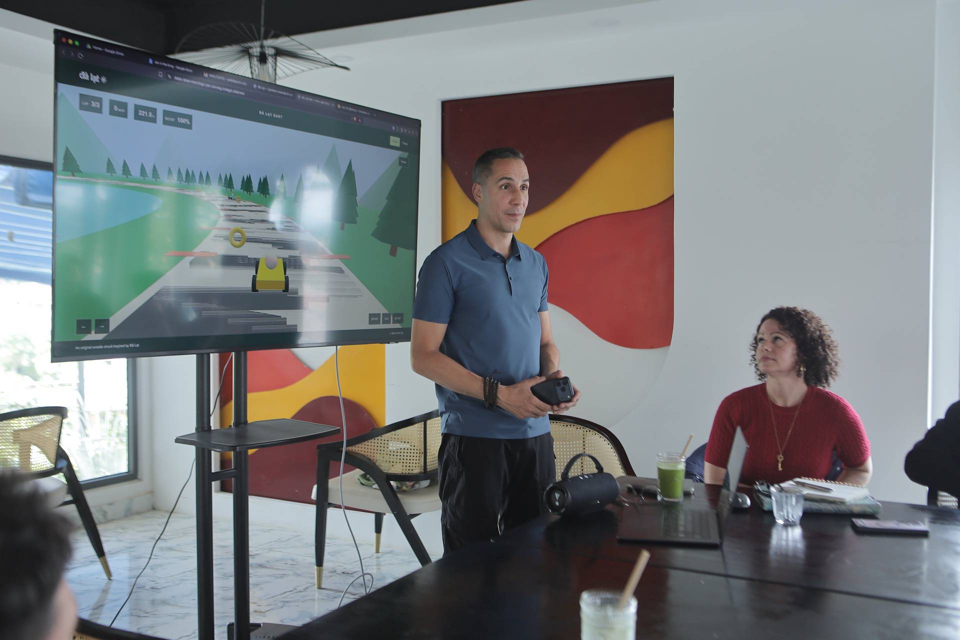 A presenter is giving a presentation in Đà Lạt, Vietnam, with a digital display showing a racing game interface. The setting appears to be a modern office or meeting room with colorful artwork on the walls.