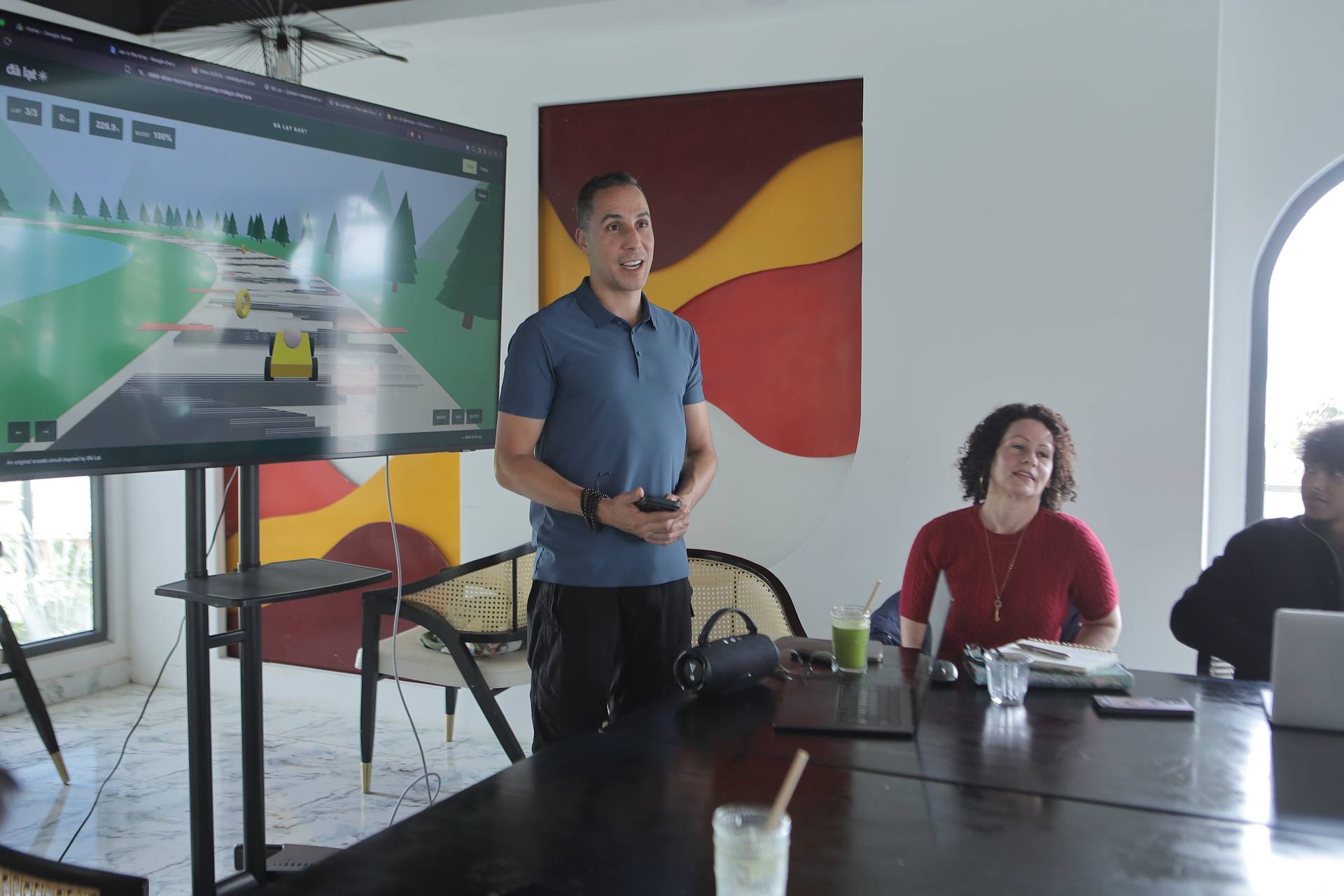 A man presents a digital game to colleagues in a modern office setting with vibrant artwork and a large screen displaying colorful graphics.