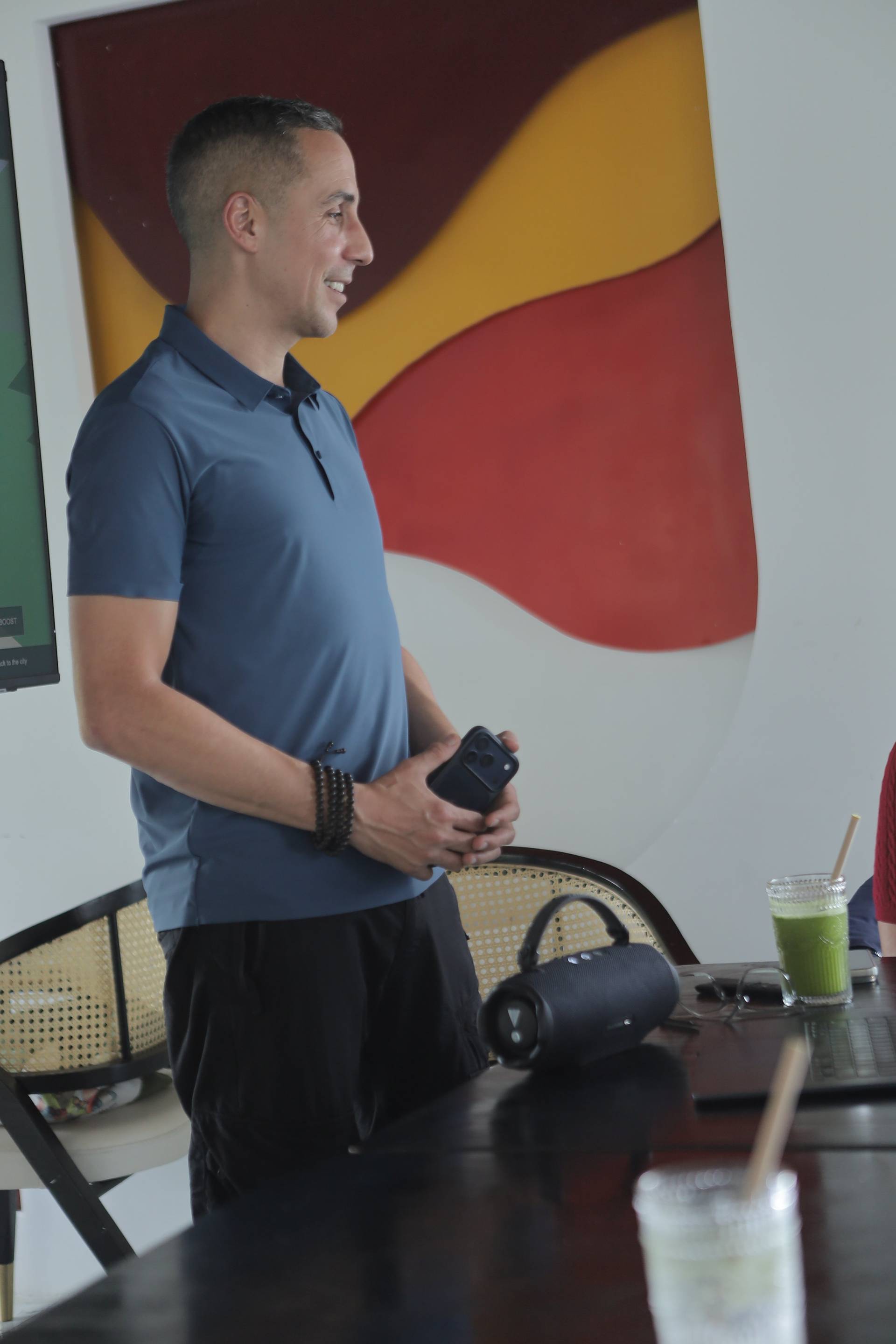 A man stands in a modern café setting in Đà Lạt, Vietnam, holding a smartphone and smiling while engaging with others around him.