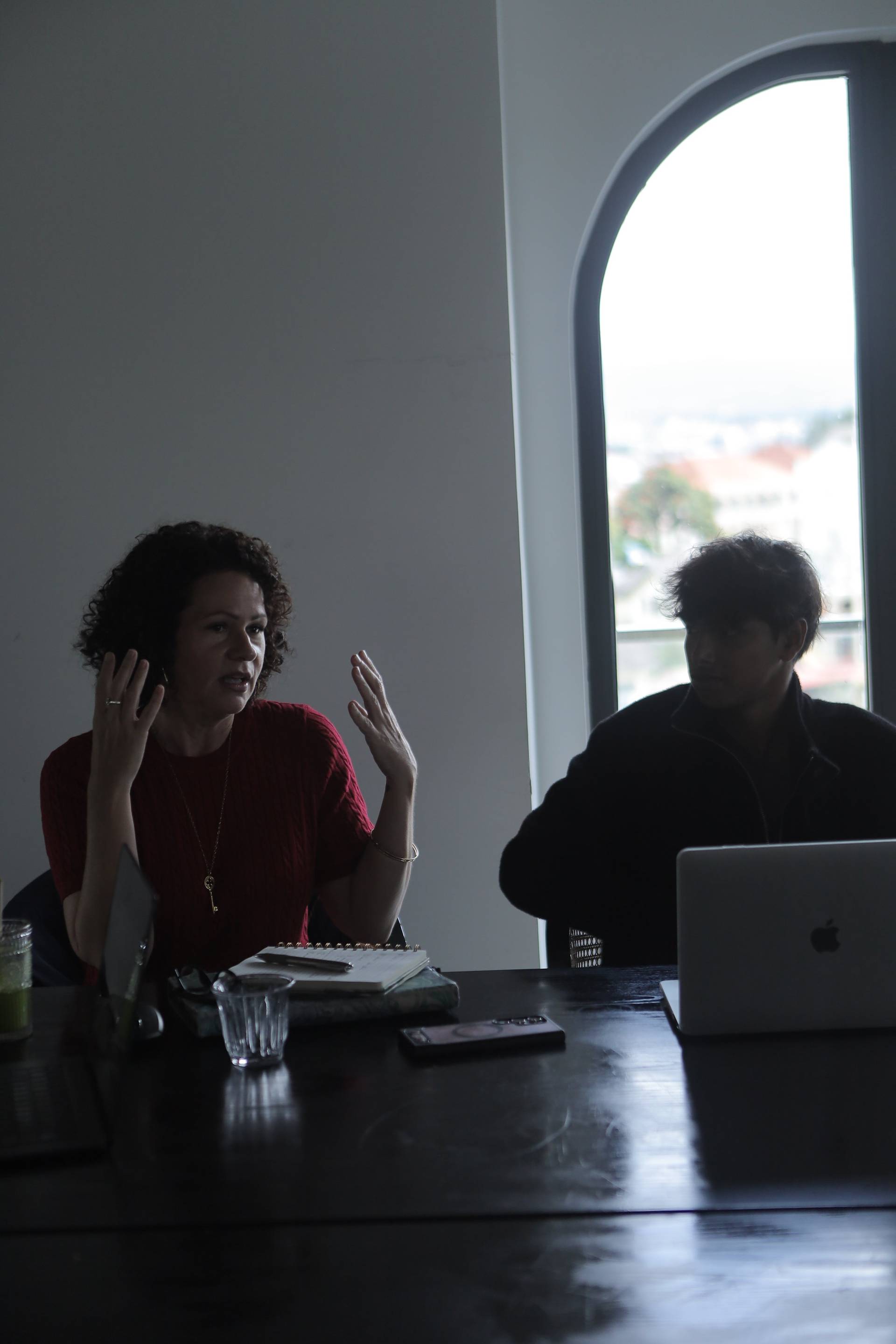 Two individuals engaged in a discussion over a table with a laptop and notebook, set against a backdrop of natural light streaming through an arched window in Đà Lạt.