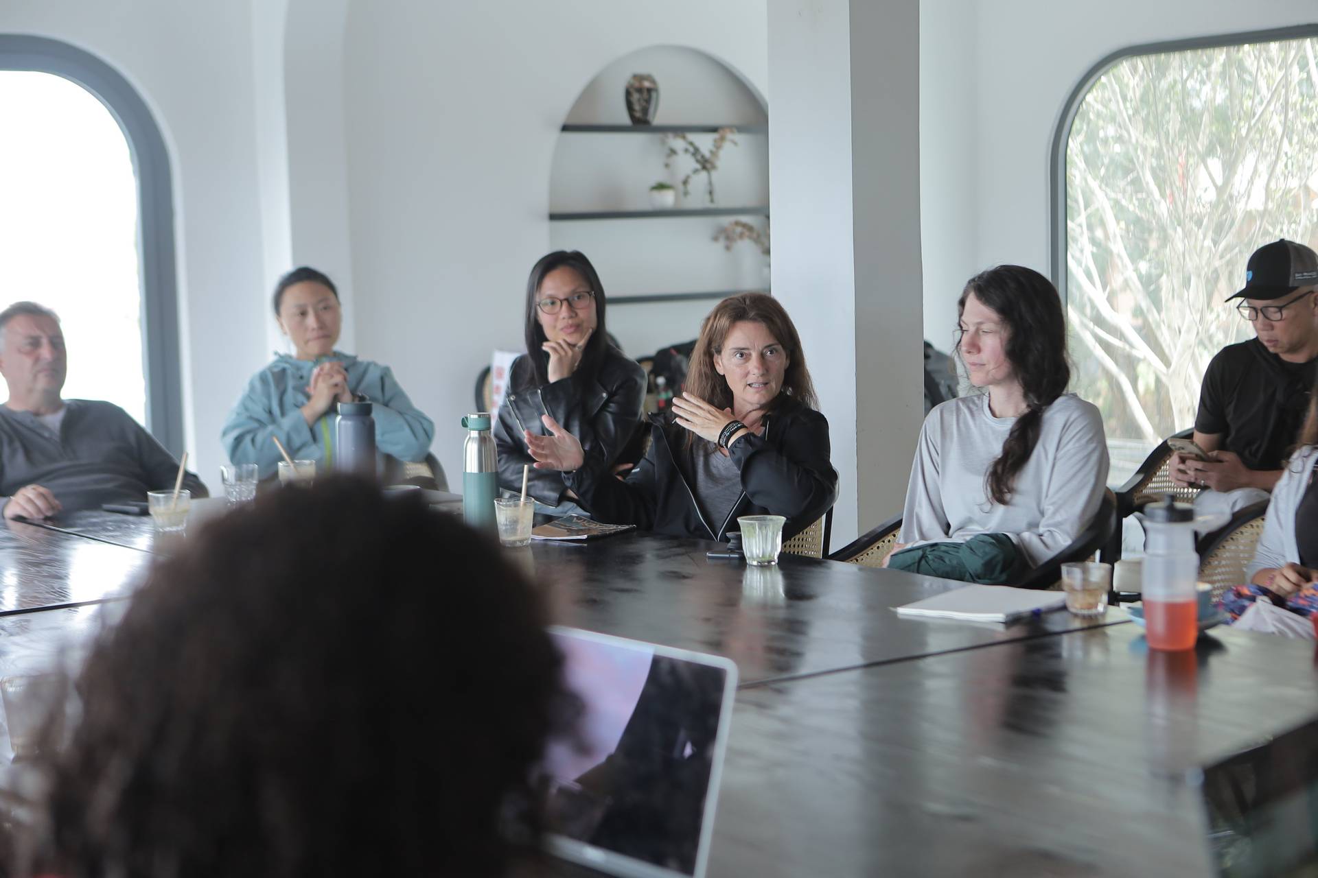 A group of individuals is engaged in a meeting or discussion around a long table in a modern, well-lit room with large windows overlooking greenery. The setting suggests a professional or collaborative environment.