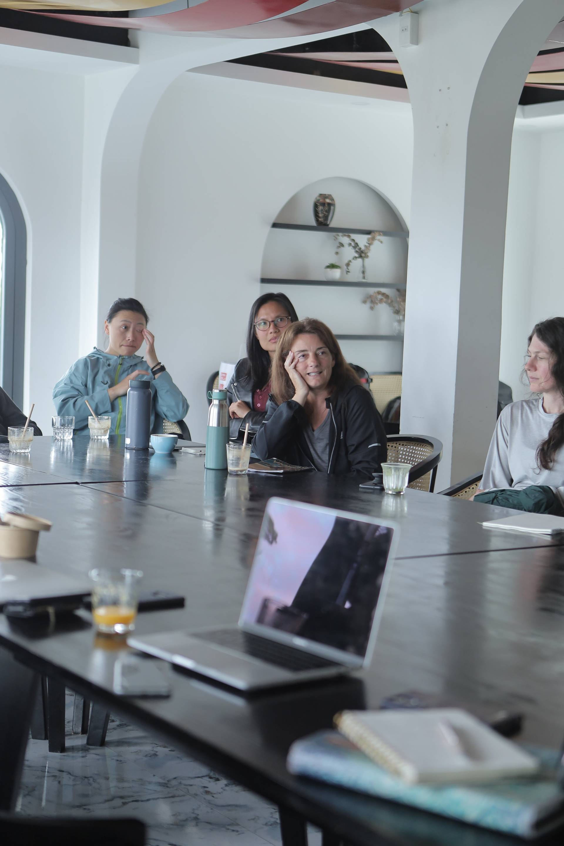 A group of individuals engaged in a meeting or discussion around a large table with laptops and refreshments, set against a modern interior backdrop.
