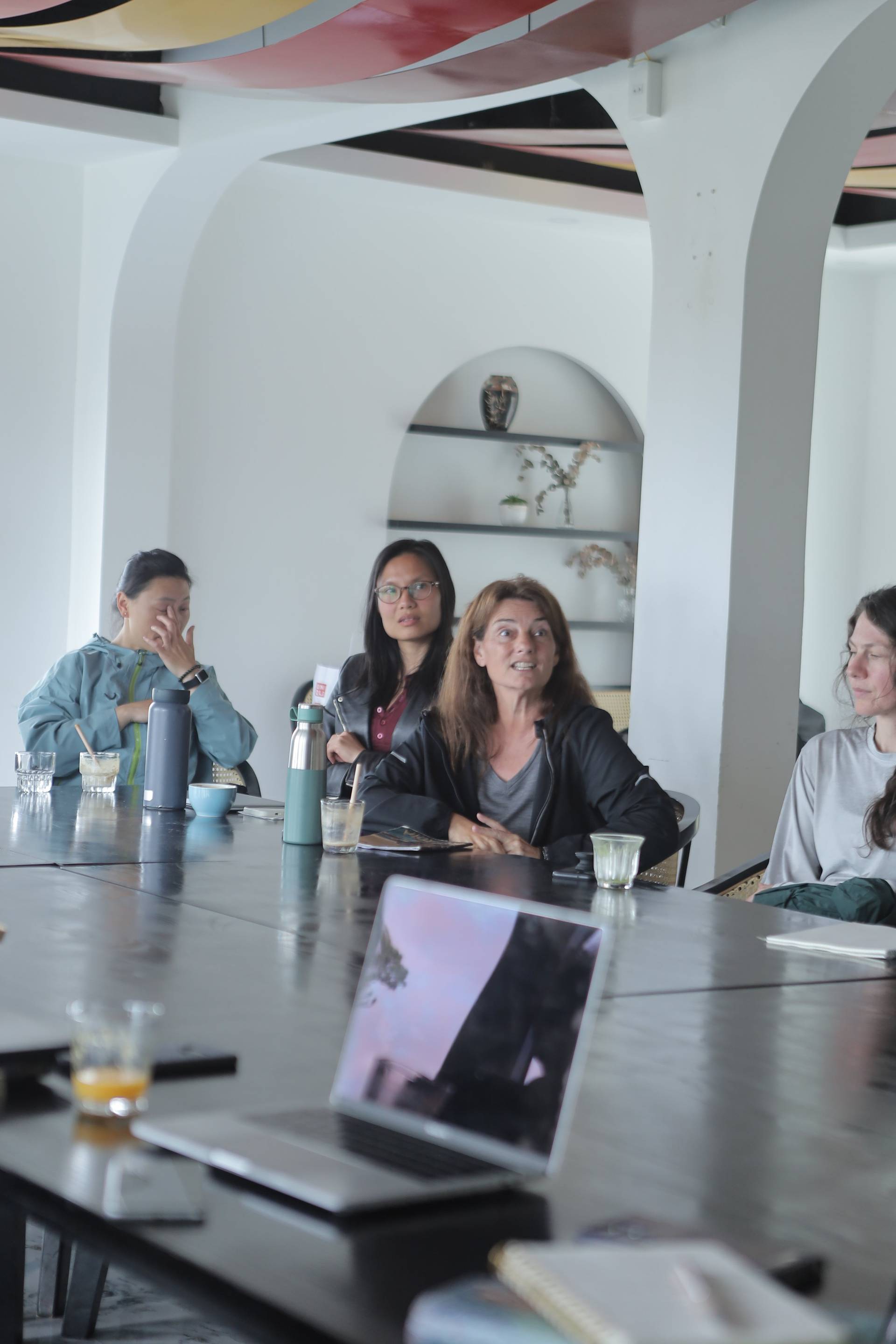 A group of individuals engaged in a meeting or discussion around a modern table setup with laptops and refreshments, set against a contemporary architectural backdrop in Đà Lạt.