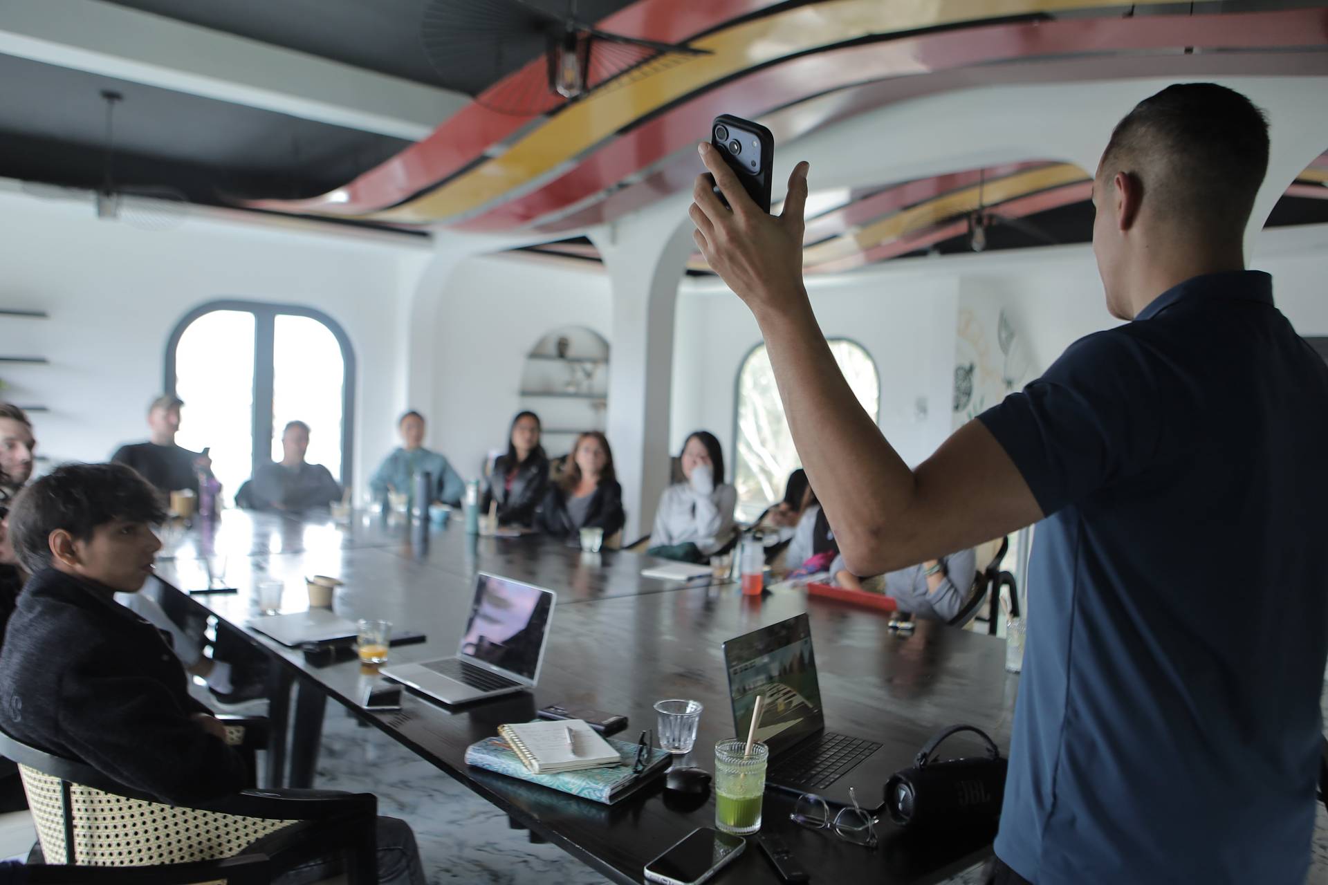 A group of people is gathered in a modern, well-lit room with large windows and artistic ceiling designs, engaging in what appears to be a presentation or meeting. One individual stands at the front holding up a smartphone, possibly recording or taking photos.