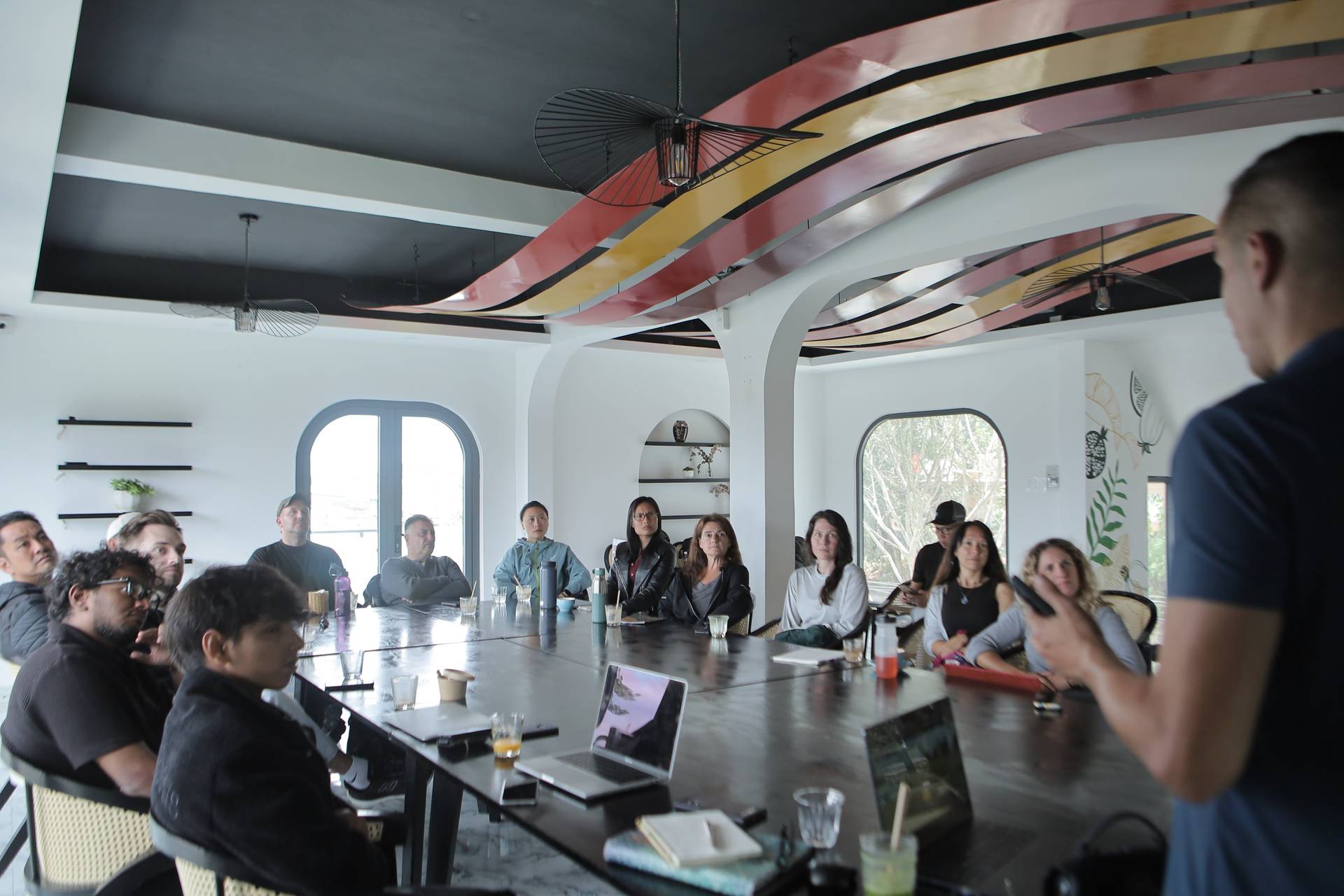 A group of professionals is engaged in a meeting around a large table in a modern conference room with vibrant ceiling art and natural light streaming through arched windows.