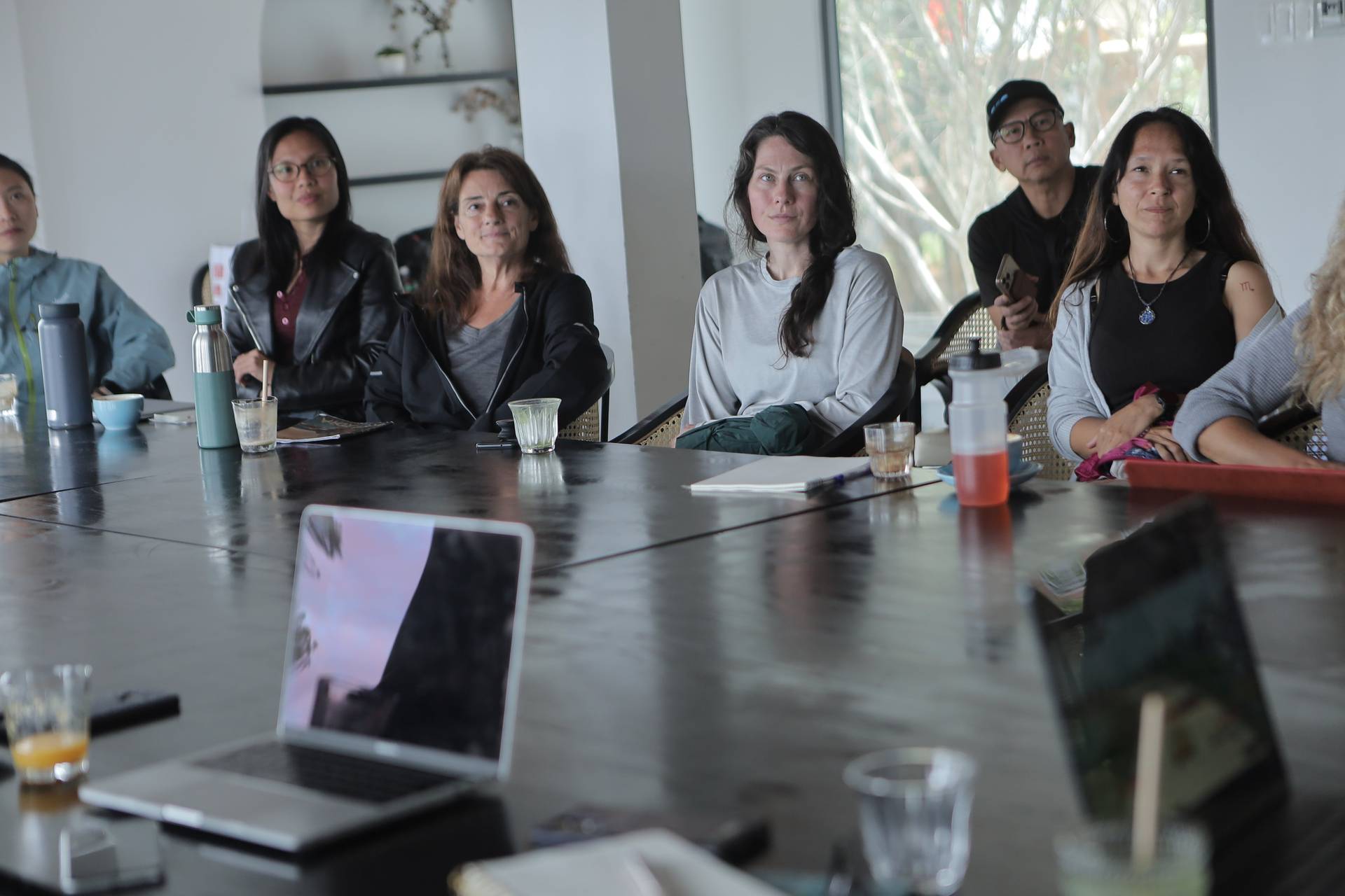 A group of people is seated around a conference table in Đà Lạt, Vietnam, engaged in what appears to be a professional or collaborative meeting with laptops and refreshments on the table.