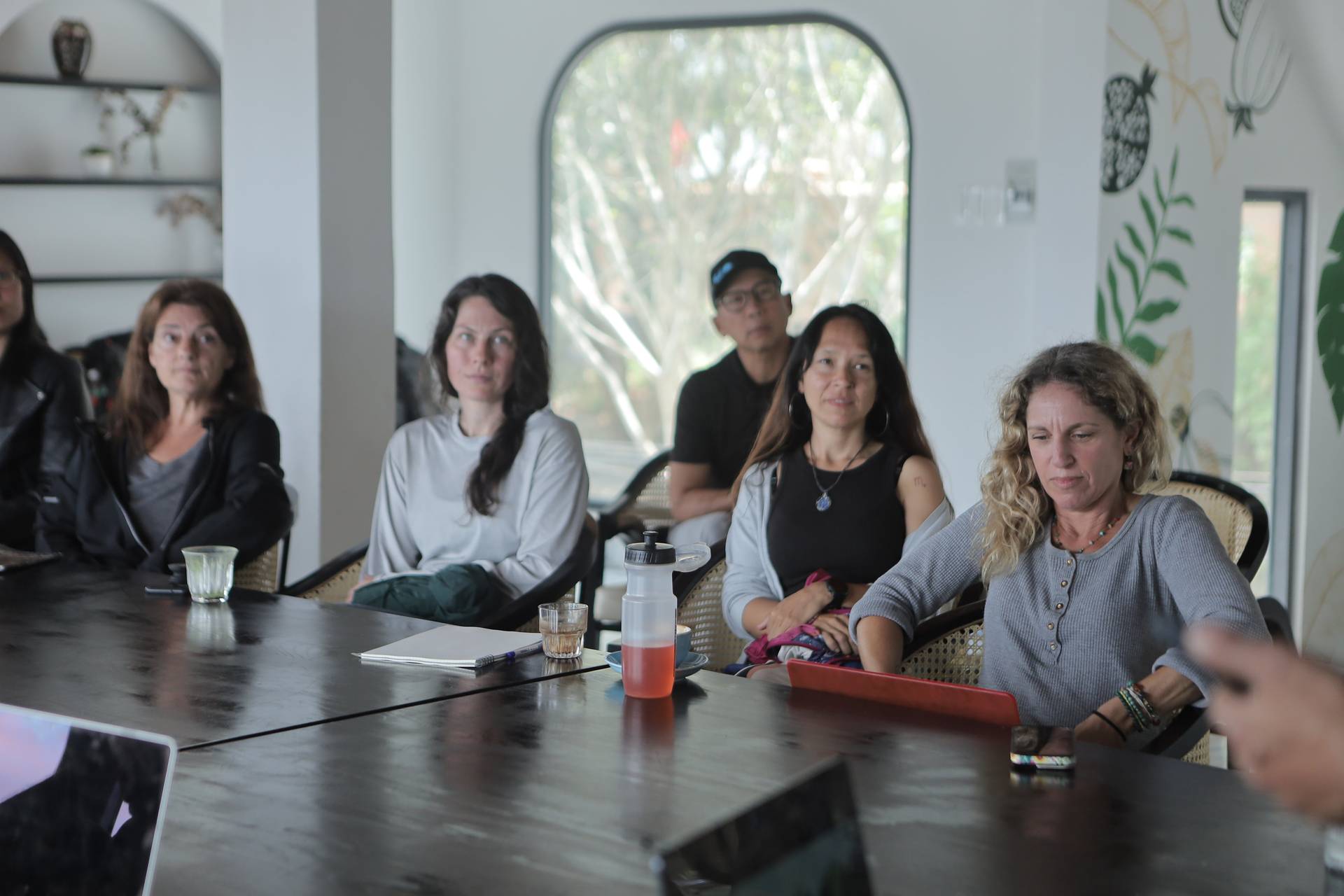 A group of people is seated around a table in a modern, well-lit room with large windows overlooking greenery, suggesting a calm and focused meeting or discussion session.