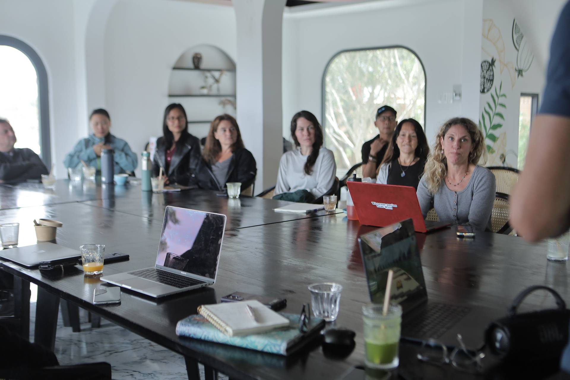 A group of professionals is engaged in a collaborative meeting around a large table in a modern, well-lit room with natural light streaming through arched windows. The setting suggests a professional or creative workspace in Đà Lạt, Vietnam.