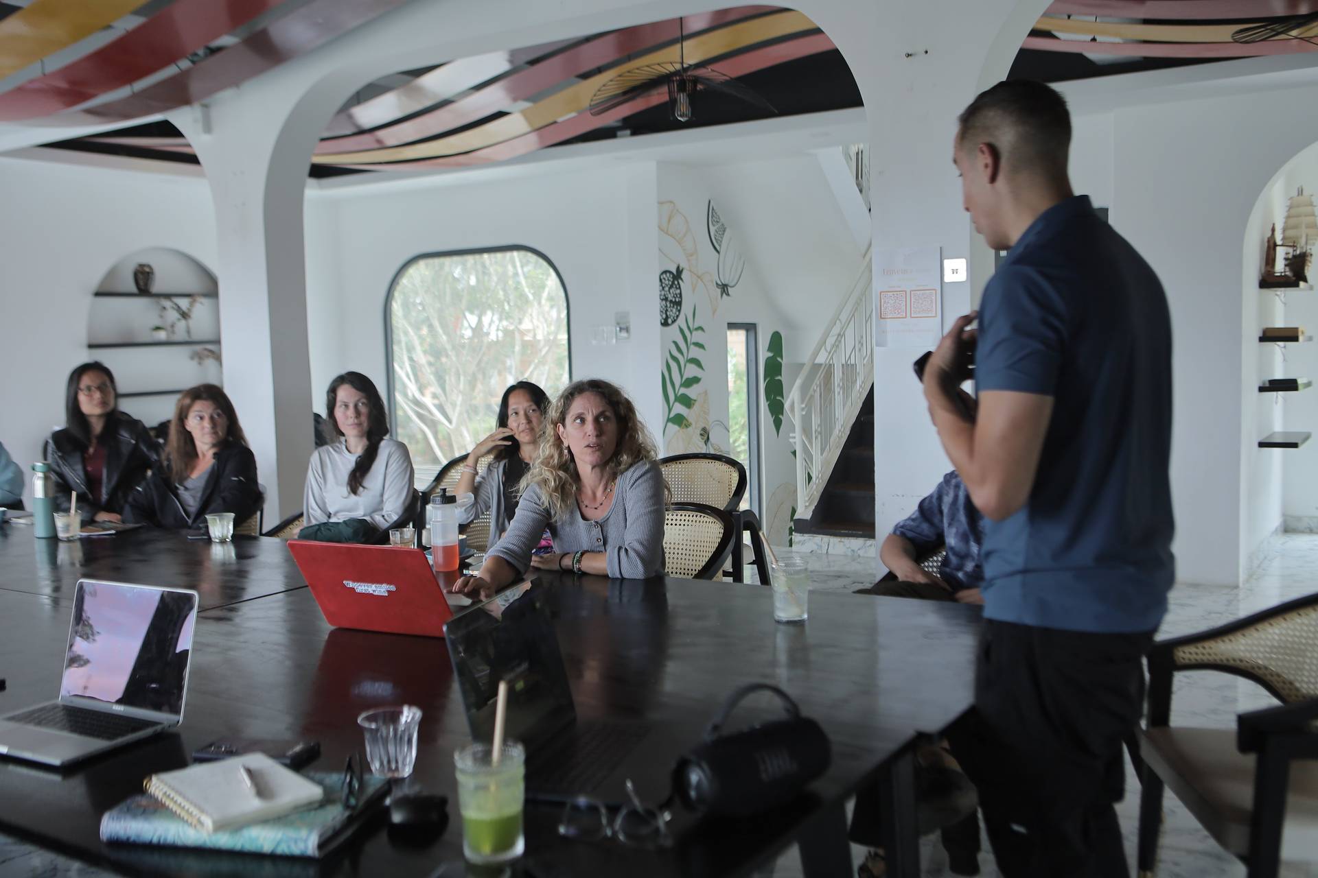 A group of individuals is gathered in a modern, well-lit room with tropical decor, engaging in what appears to be an educational or professional discussion led by someone standing at the front holding a presentation board.