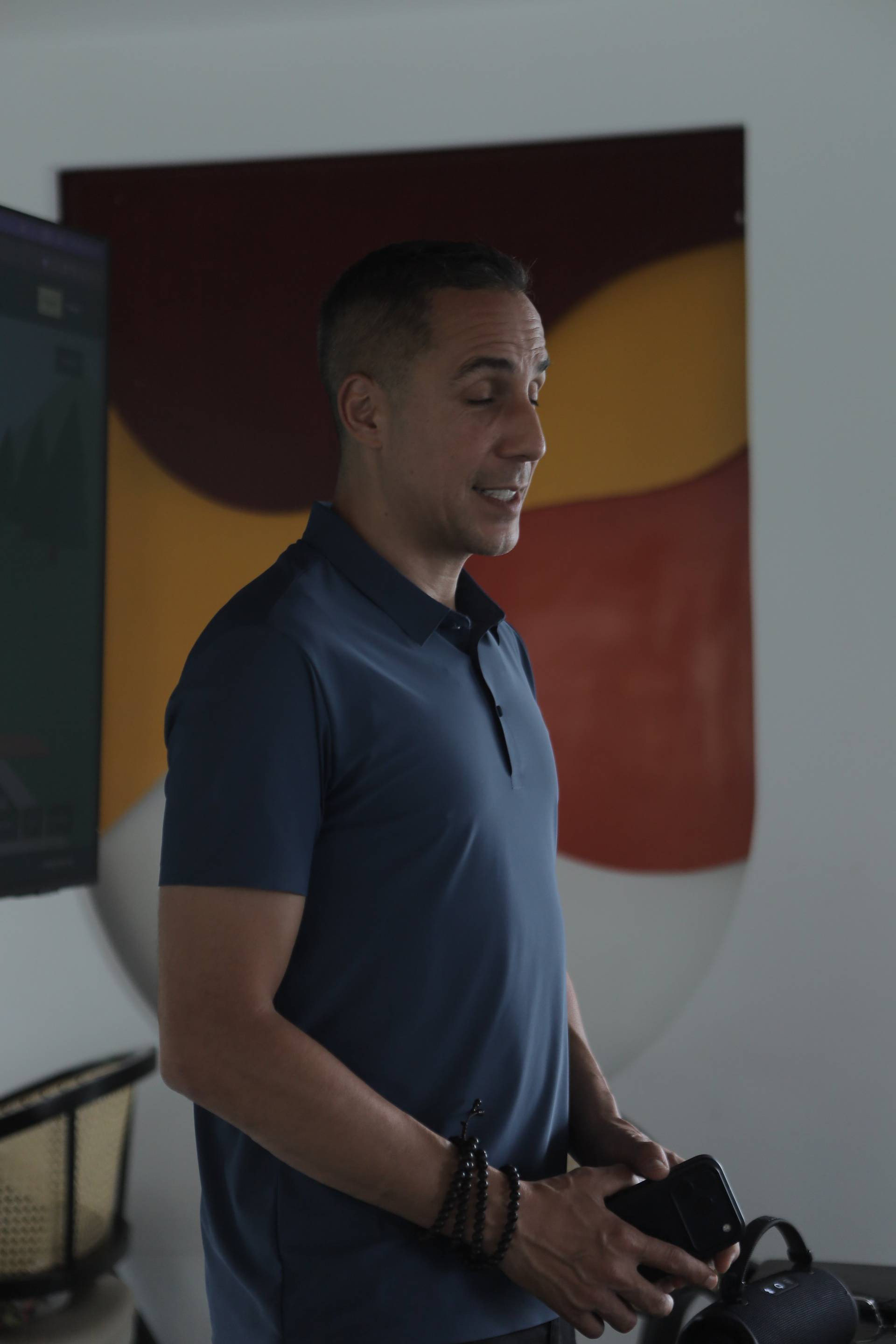 A man in a blue polo shirt stands indoors with a colorful abstract painting behind him, holding a smartphone and appearing to be engaged in conversation or presentation.