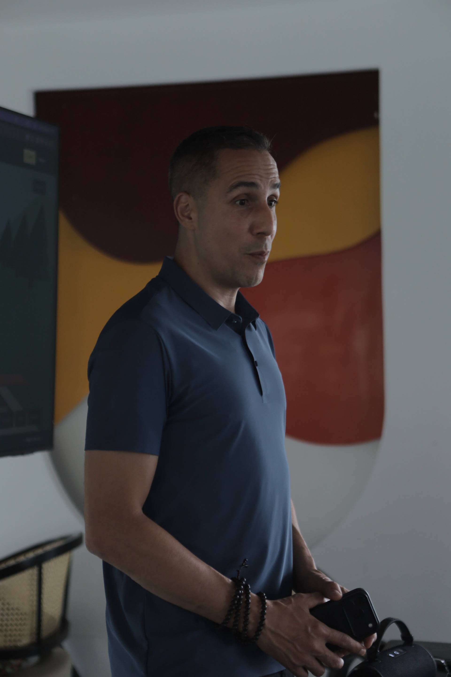 A man in a dark blue polo shirt stands indoors with a modern abstract painting behind him, holding a smartphone and a small speaker, suggesting a casual tech-focused event.
