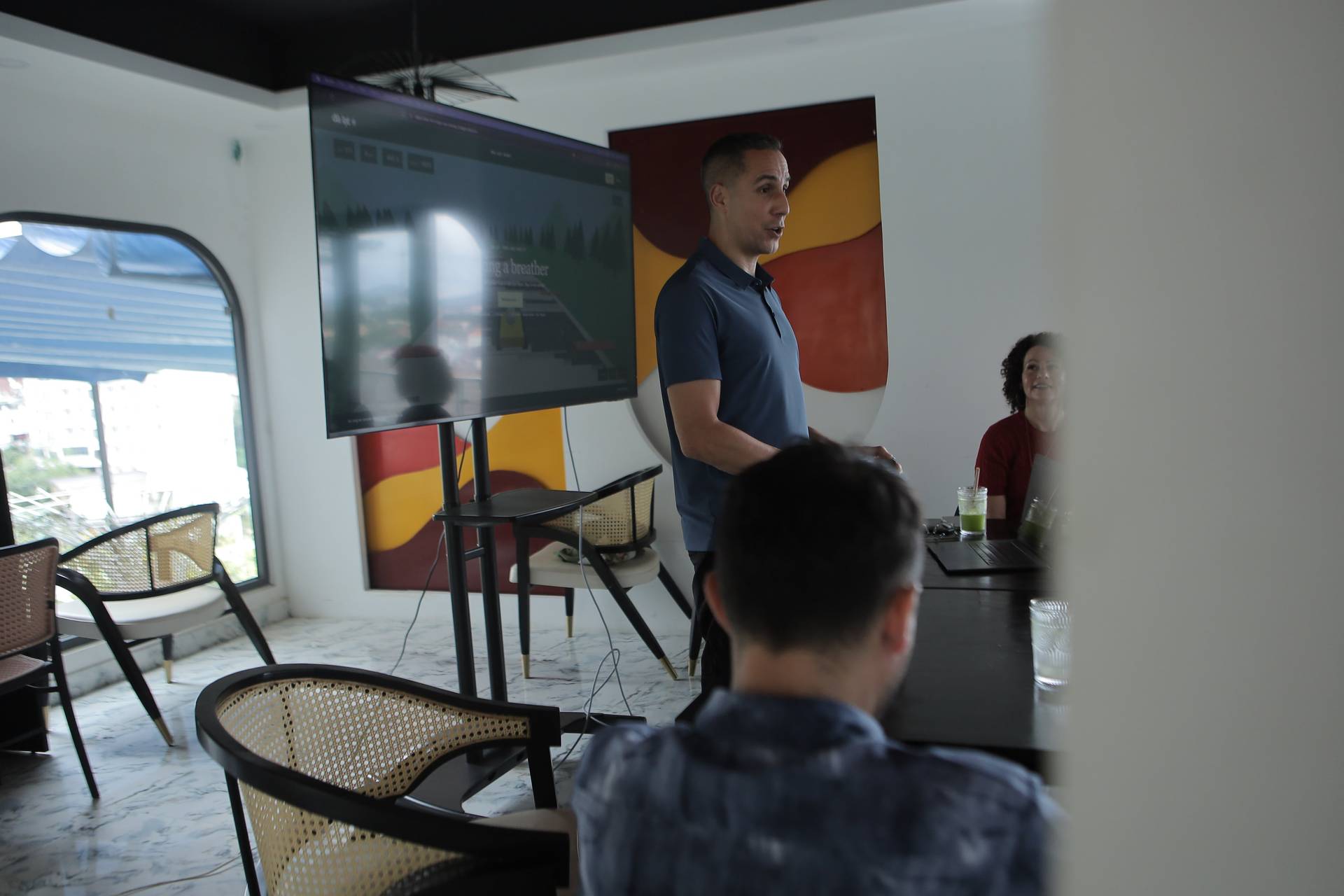 A man presents in front of a large screen to an audience seated around a table with drinks and laptops, set against a backdrop of colorful artwork and a view of Đà Lạt's urban landscape.