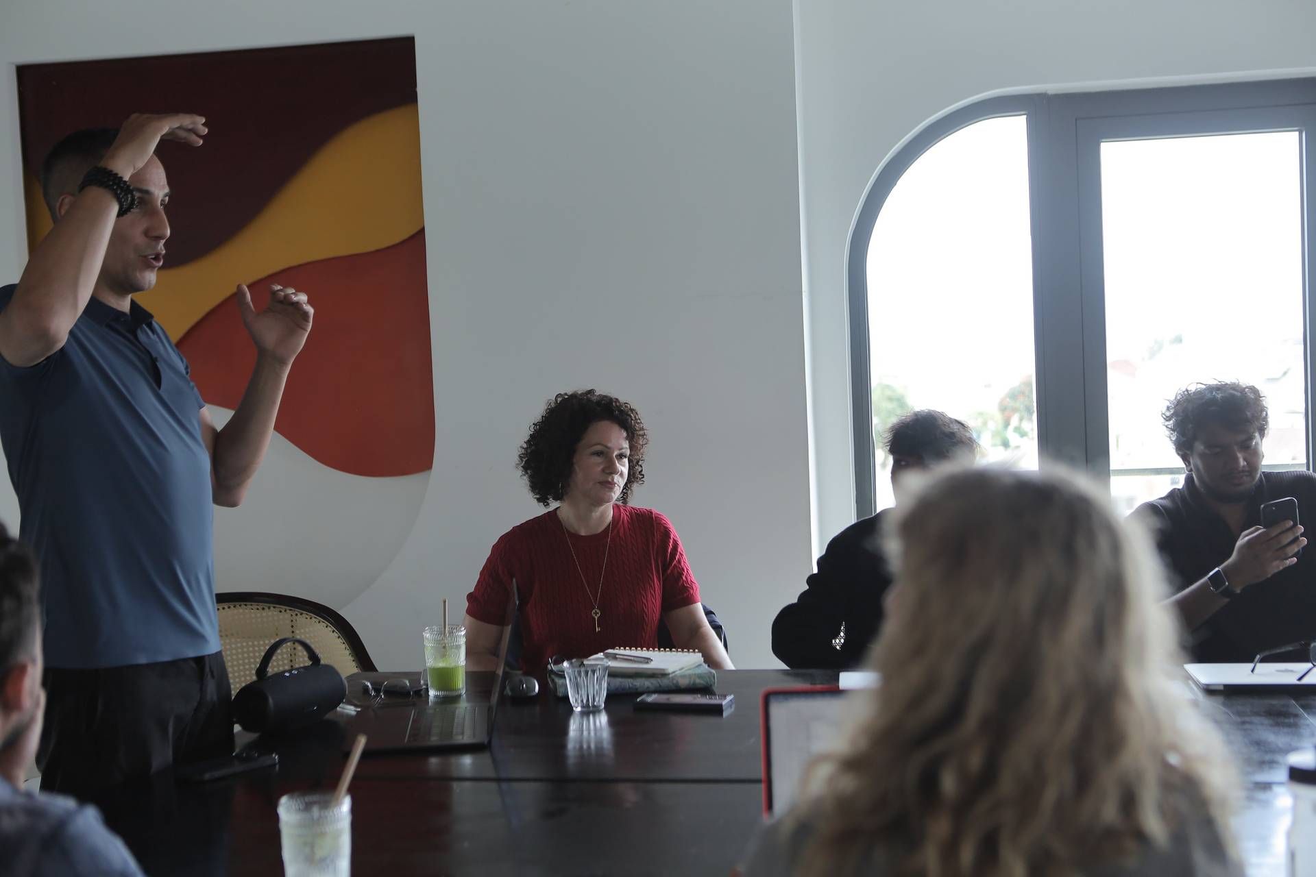 A group of people is engaged in a meeting or presentation in a modern office setting with large windows and vibrant artwork. The atmosphere appears professional yet relaxed.