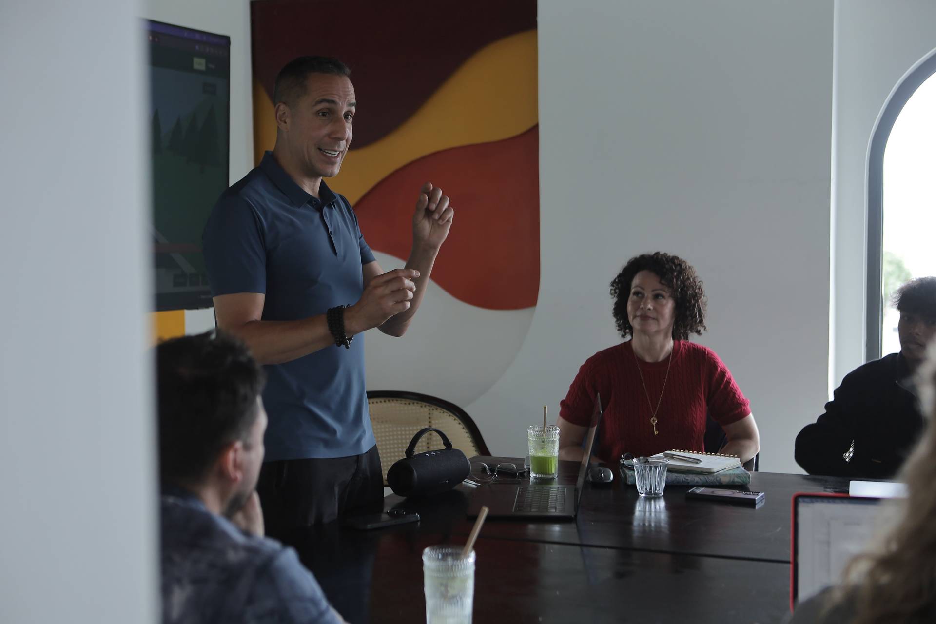 A man stands addressing a group seated around a table in a modern office setting with vibrant artwork in the background, suggesting a professional or collaborative meeting.