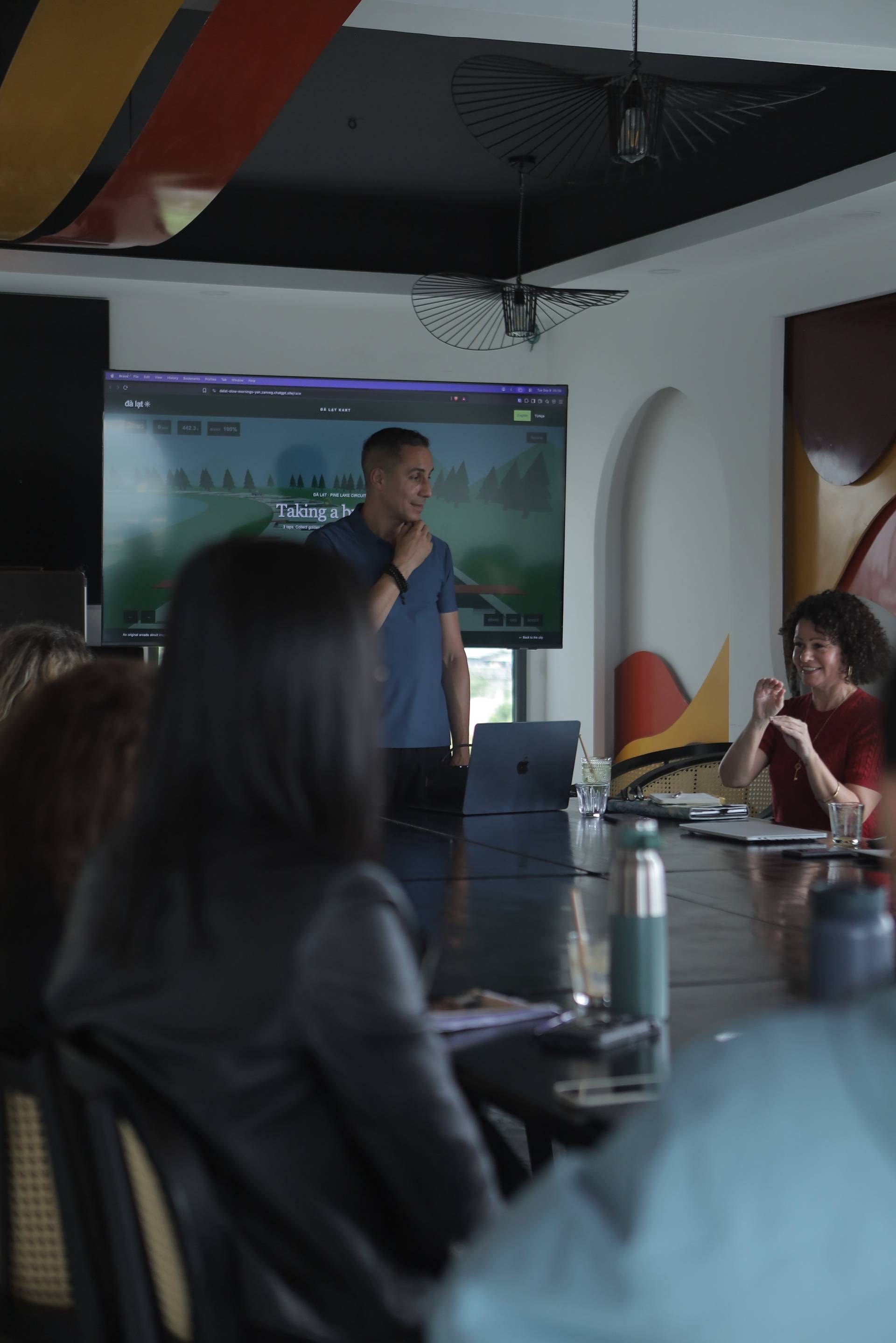 A group of people is engaged in a presentation or meeting in a modern conference room with a large screen displaying a scenic image from Đà Lạt, Vietnam.