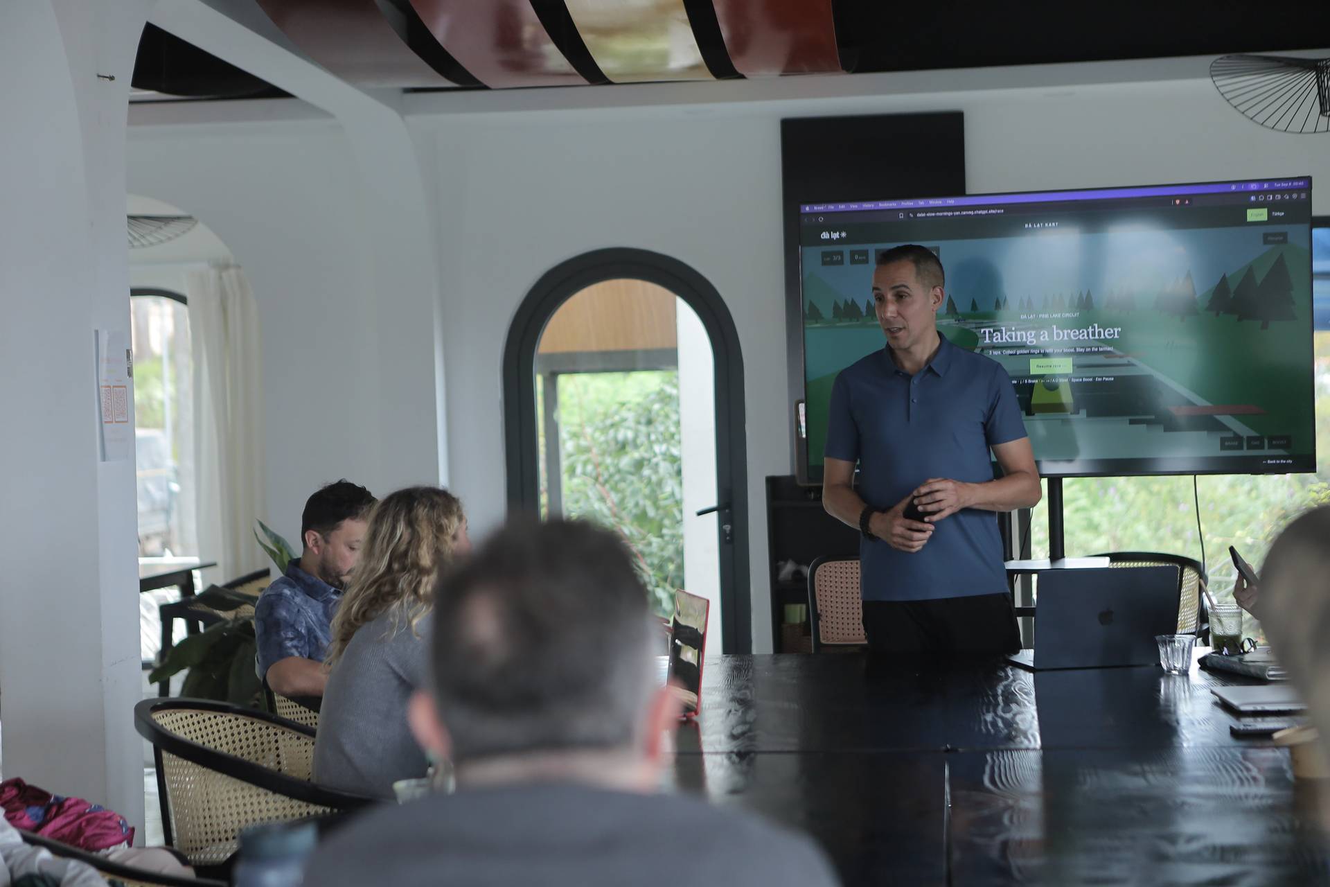 A man presents a digital game to a group of attendees in a modern conference room with large windows overlooking greenery in Đà Lạt, Vietnam.