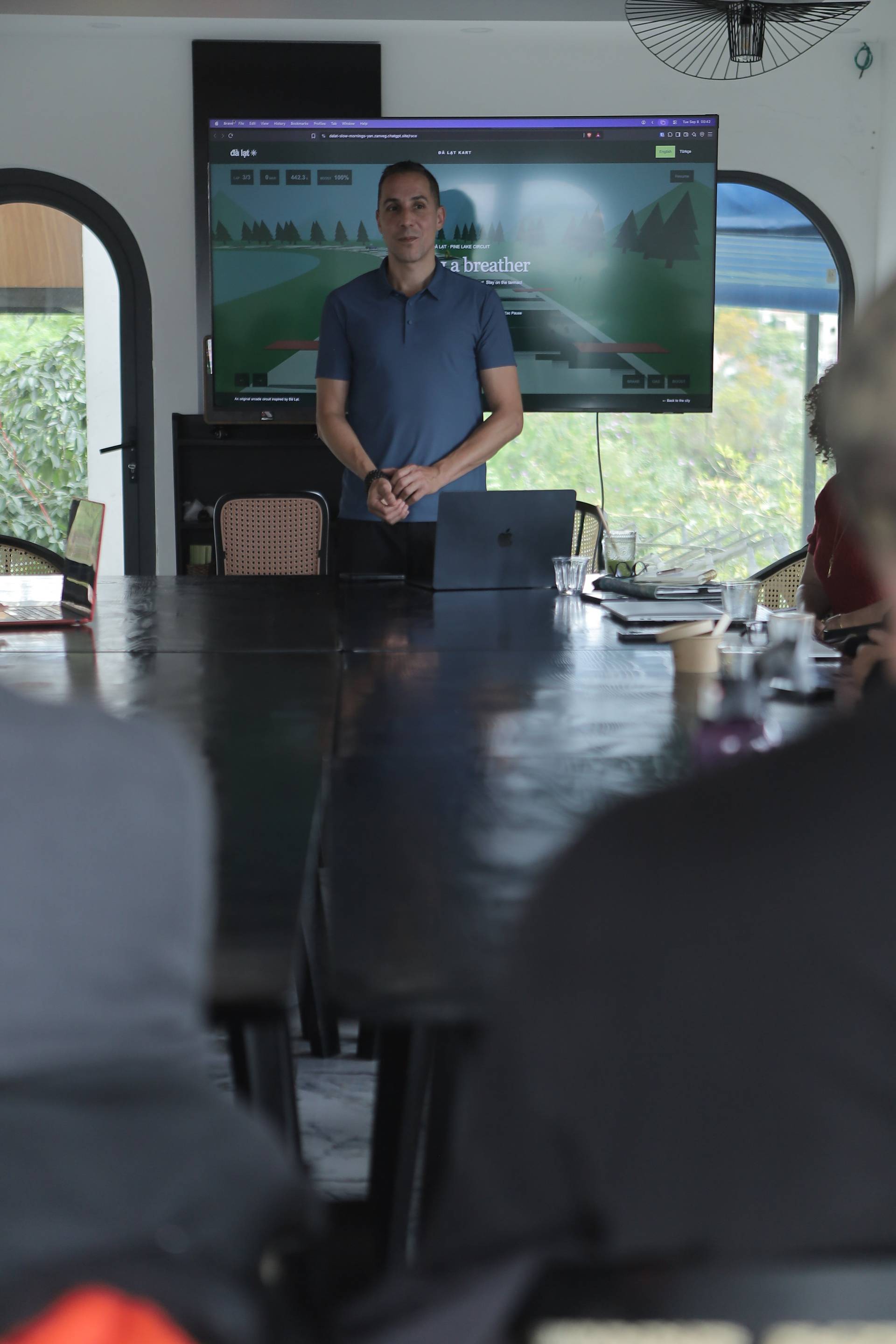 A professional presentation in a modern conference room with a large screen displaying a racing game interface, set against the backdrop of Đà Lạt's scenic landscape.