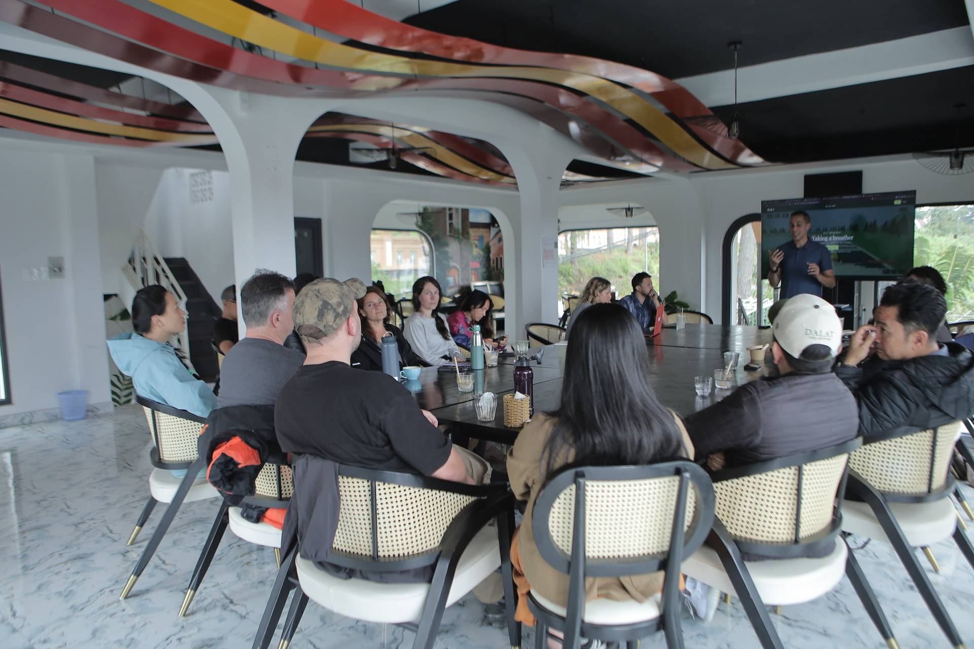 A group of people is gathered around a large table in a modern, open-air setting in Đà Lạt, Vietnam, listening to a presentation by someone standing at the front.