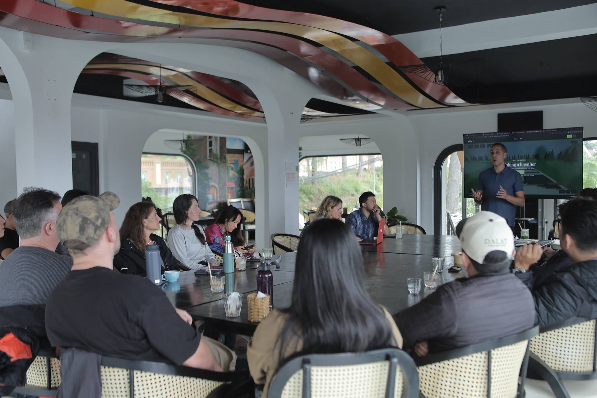 A group of people is attentively listening to a presentation in a modern café setting with a scenic view of Đà Lạt's lush greenery.