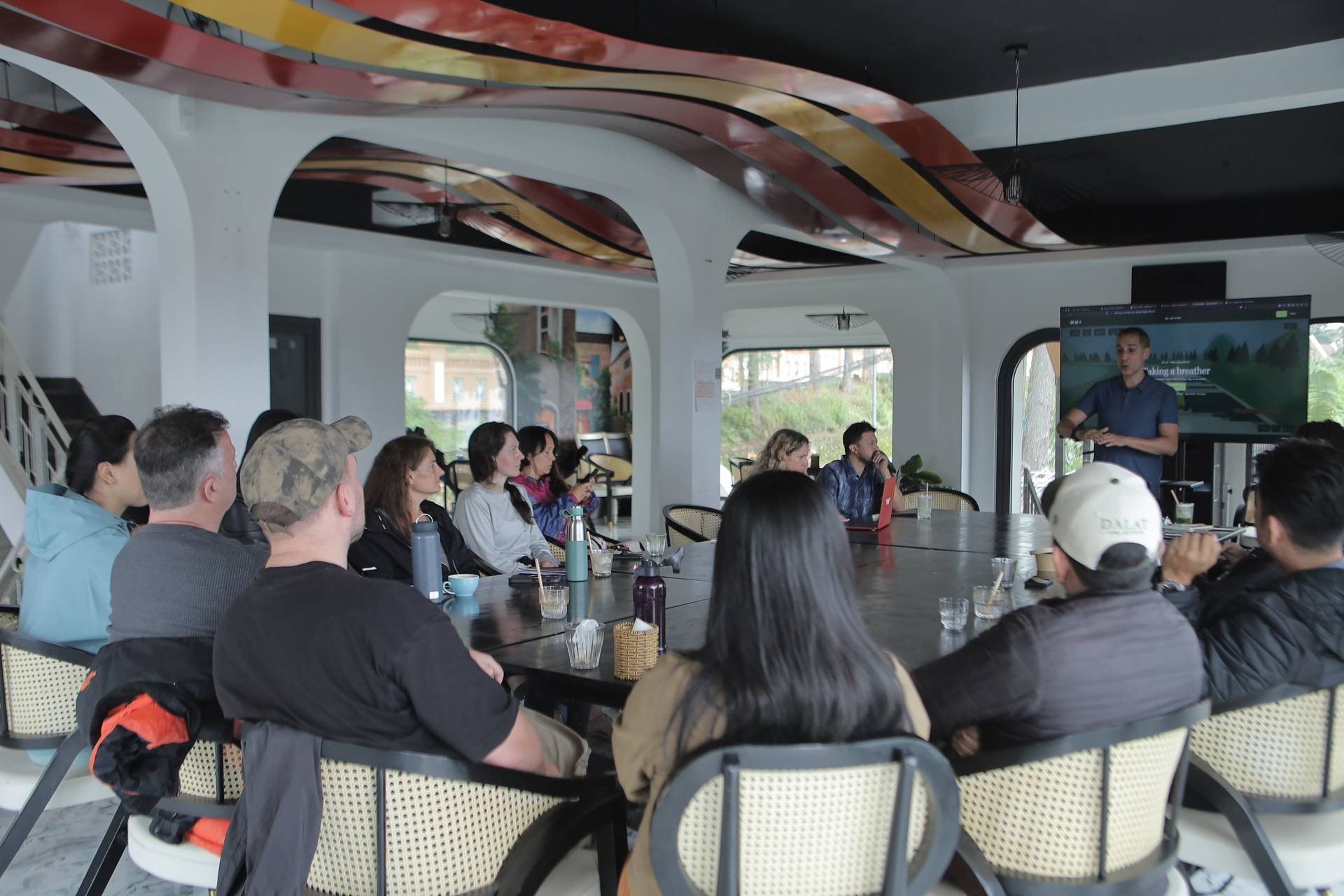 A group of people is gathered around a large table in a modern, open-air cafe in Đà Lạt, Vietnam, listening to a presentation by someone standing at the front.