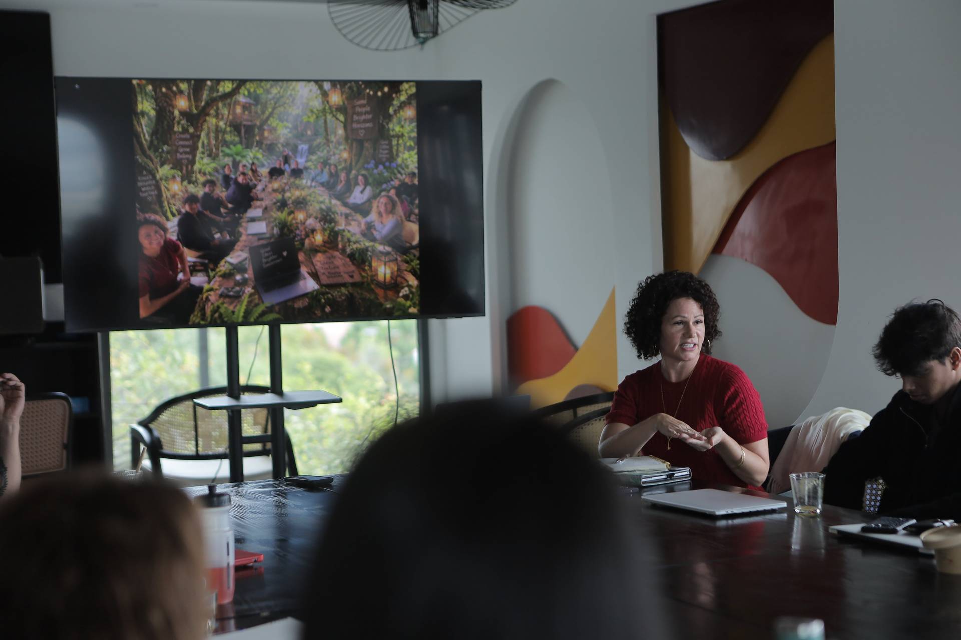 A woman in a red sweater leads a discussion or presentation to a group of attendees seated around a table in a modern conference room with large windows overlooking lush greenery.