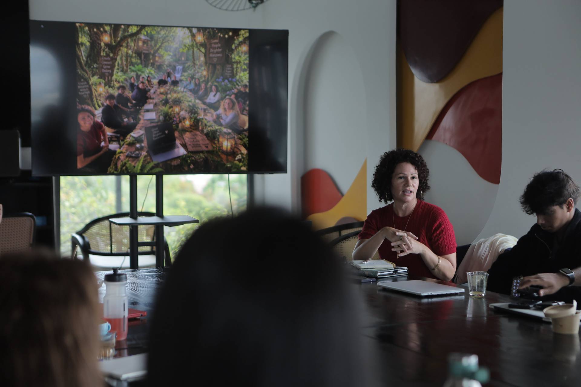 A woman in a red dress leads a discussion during a meeting in Đà Lạt, Vietnam, with a large screen displaying an image of people gathered outdoors in a lush setting.
