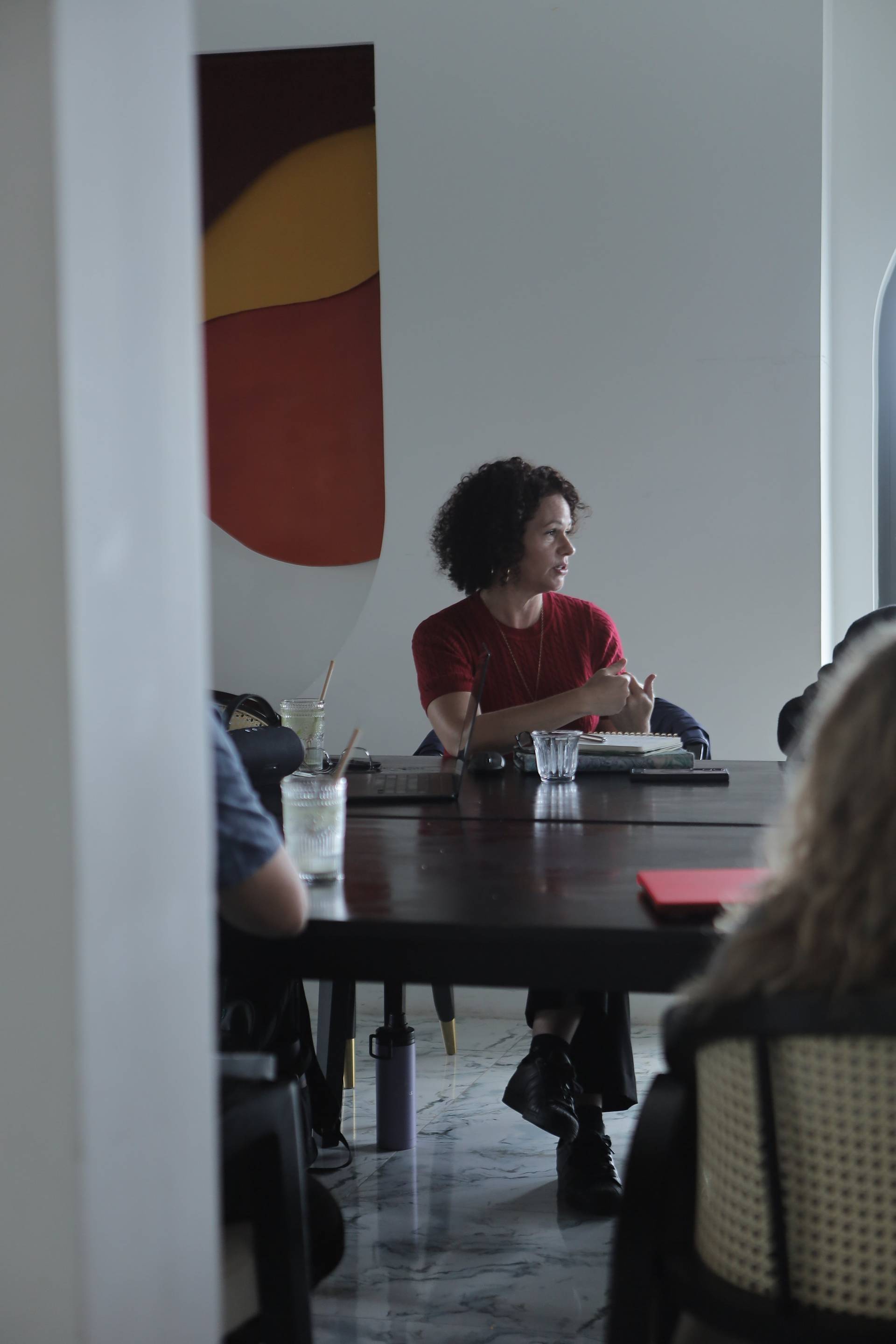 A group of people engaged in a casual meeting or discussion around a table in a modern café setting with vibrant artwork in the background.