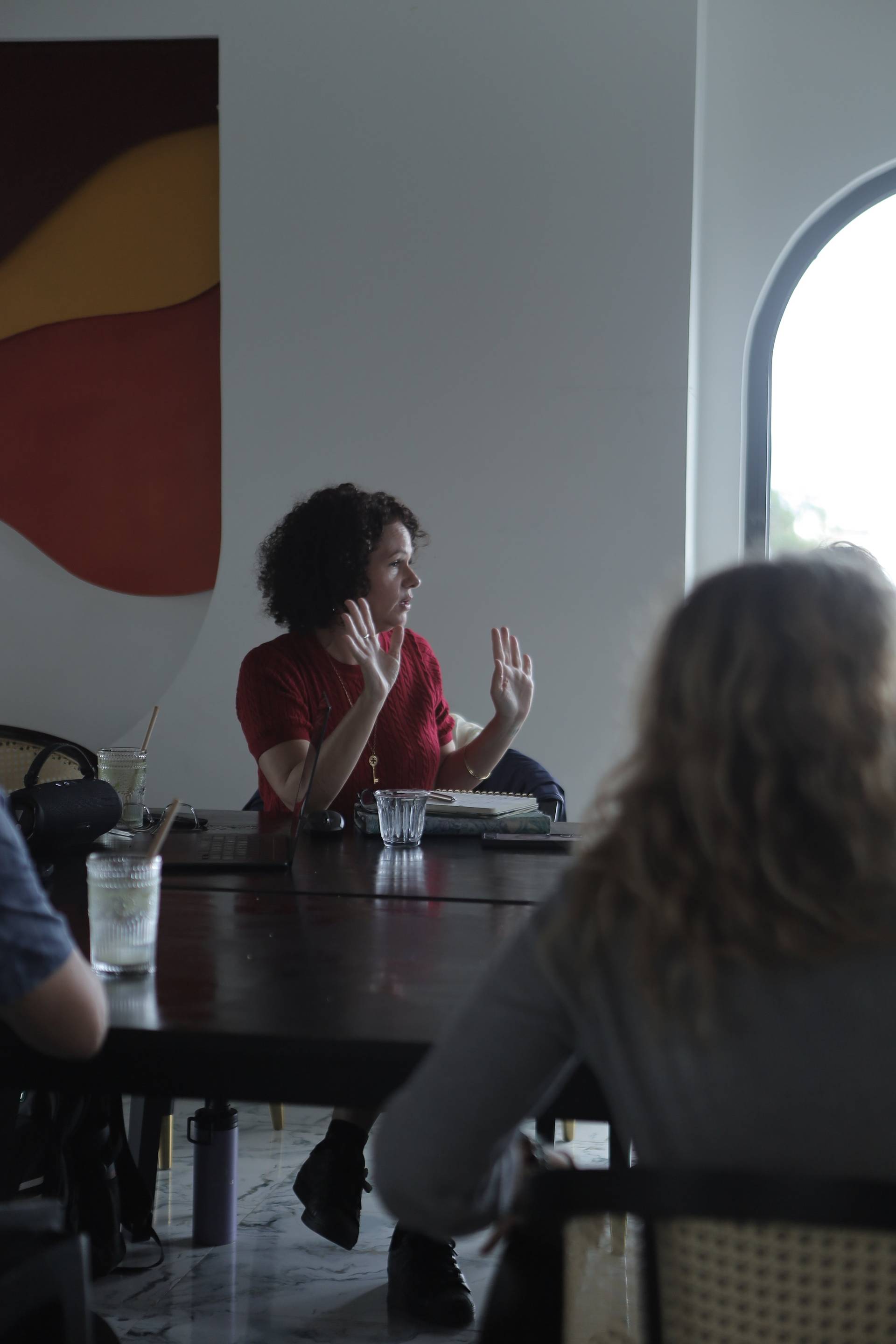 A woman in a red sweater gestures animatedly during a meeting or discussion in a modern café setting with large windows and colorful wall art.