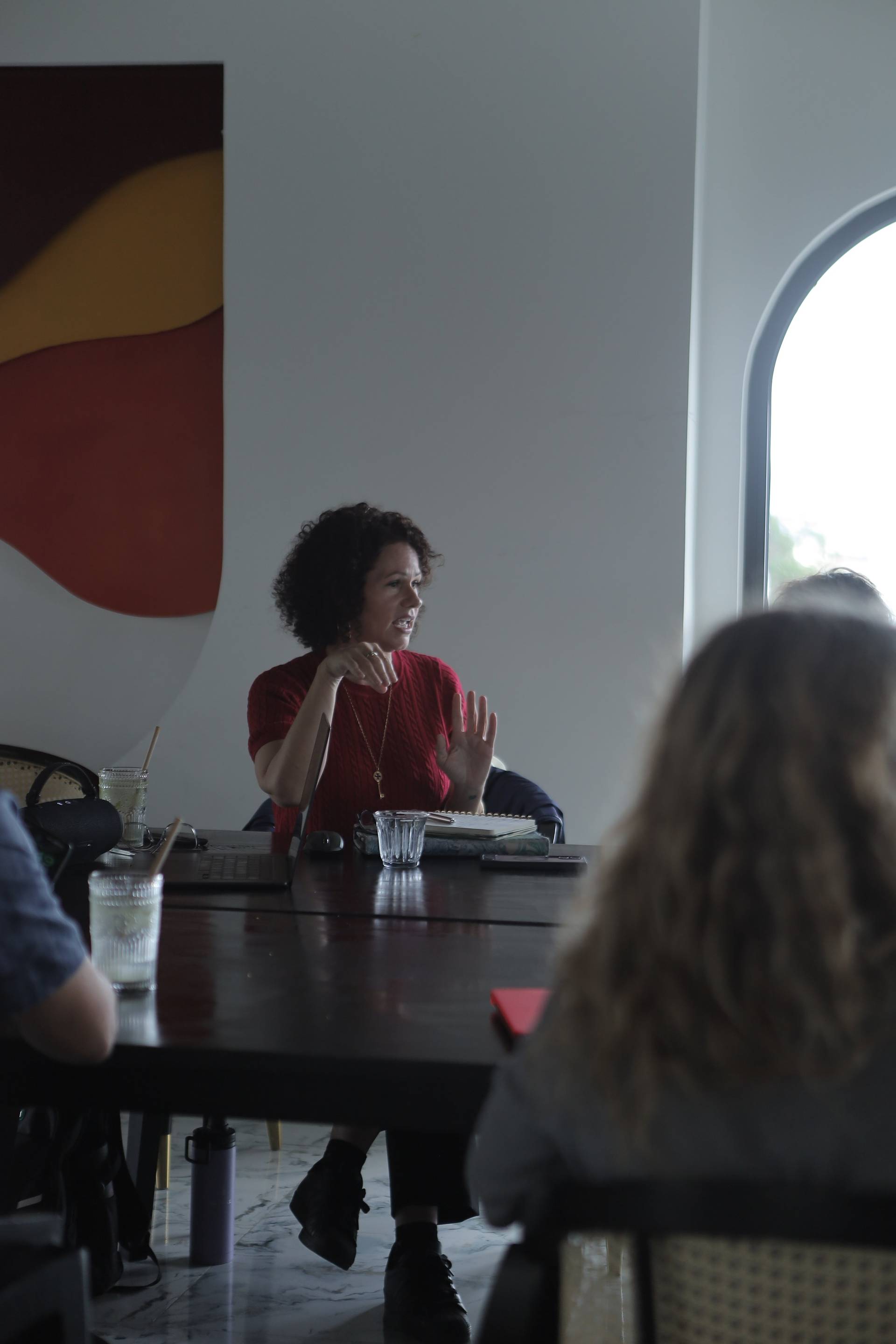 A group of people engaged in a lively discussion around a table in a modern café setting with vibrant wall art and natural light streaming through an arched window.