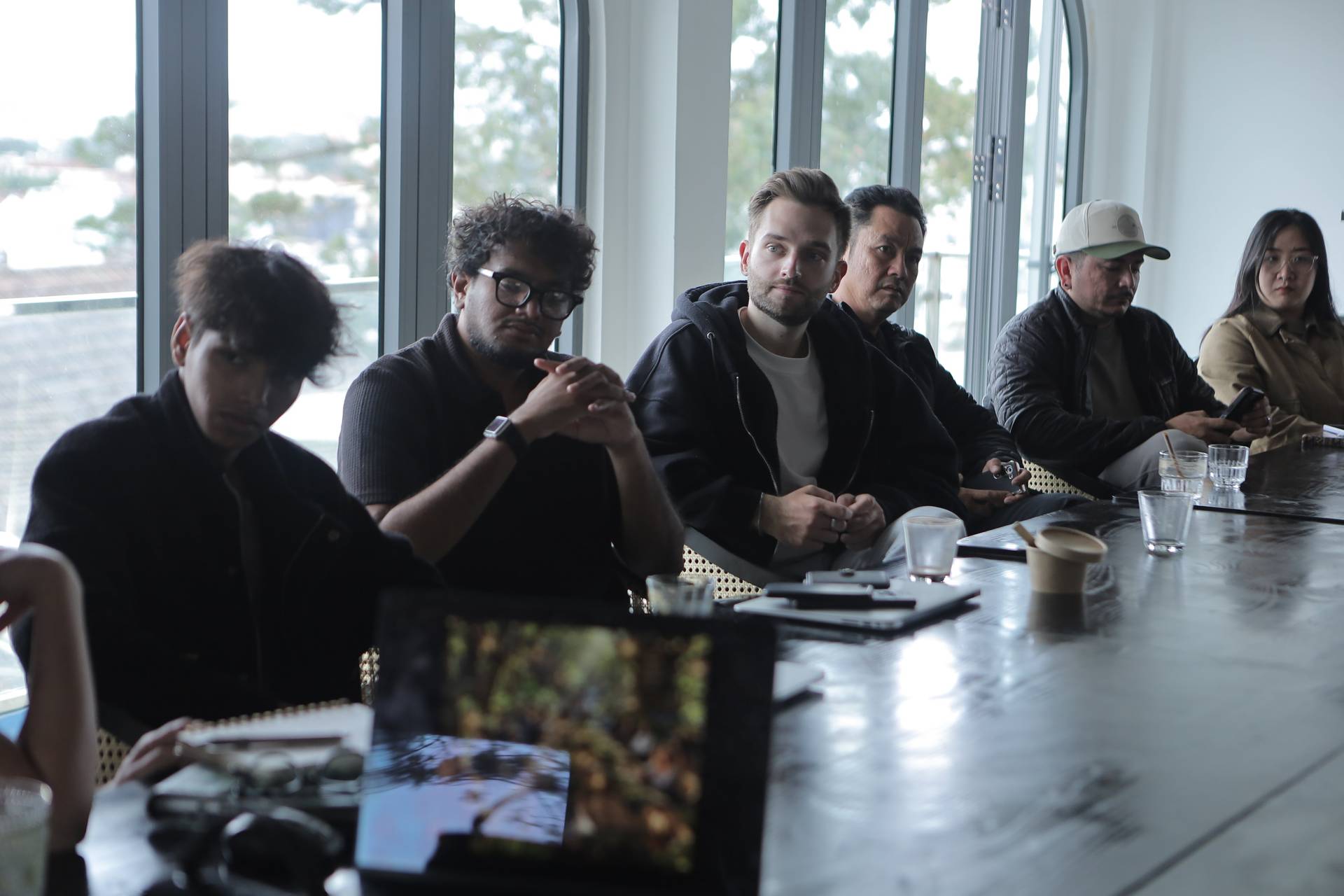 A group of individuals engaged in a meeting or discussion around a long table with modern decor and large windows overlooking a scenic view.