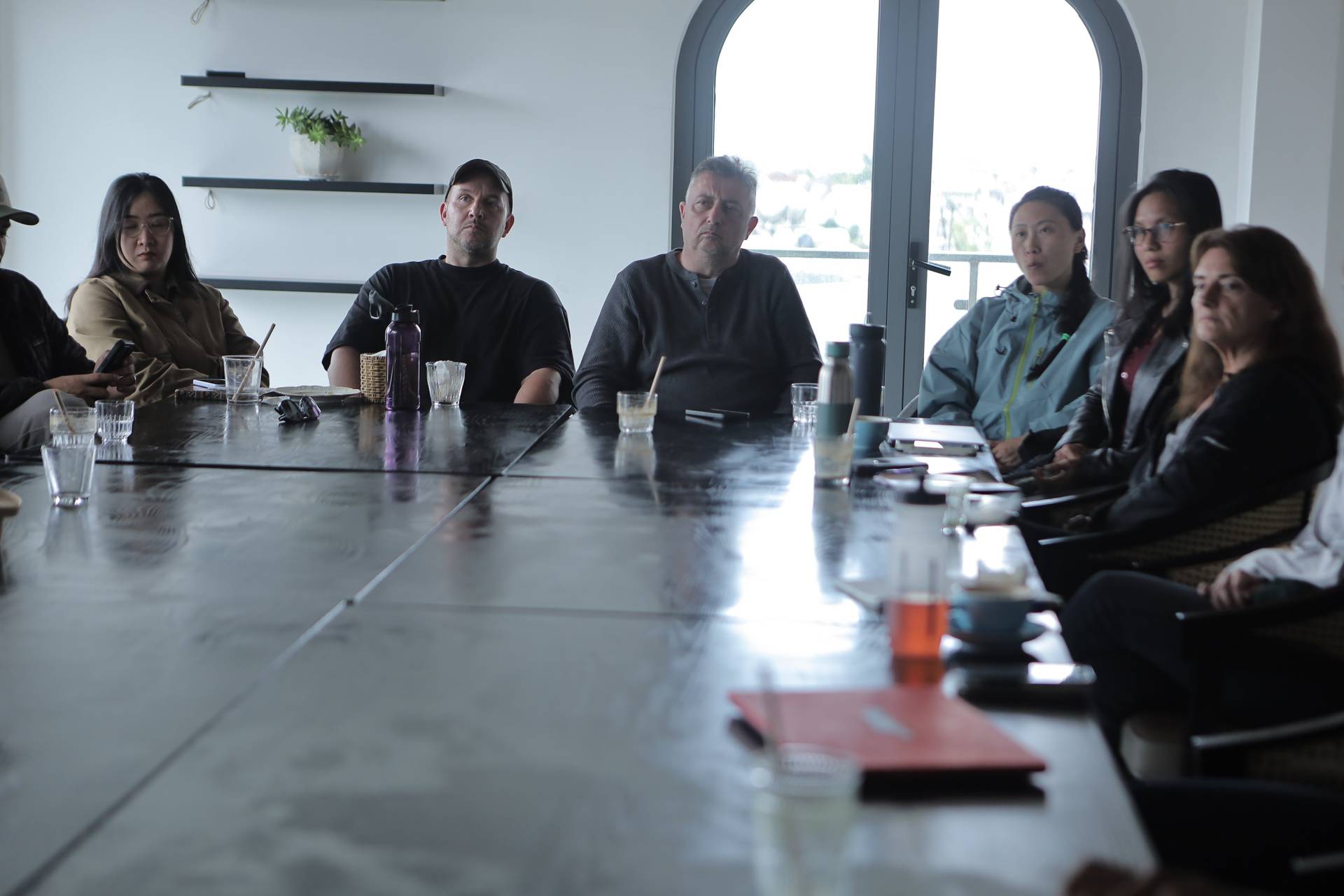 A group of individuals is seated around a long table in a modern setting with large windows, engaged in what appears to be a serious discussion or meeting.