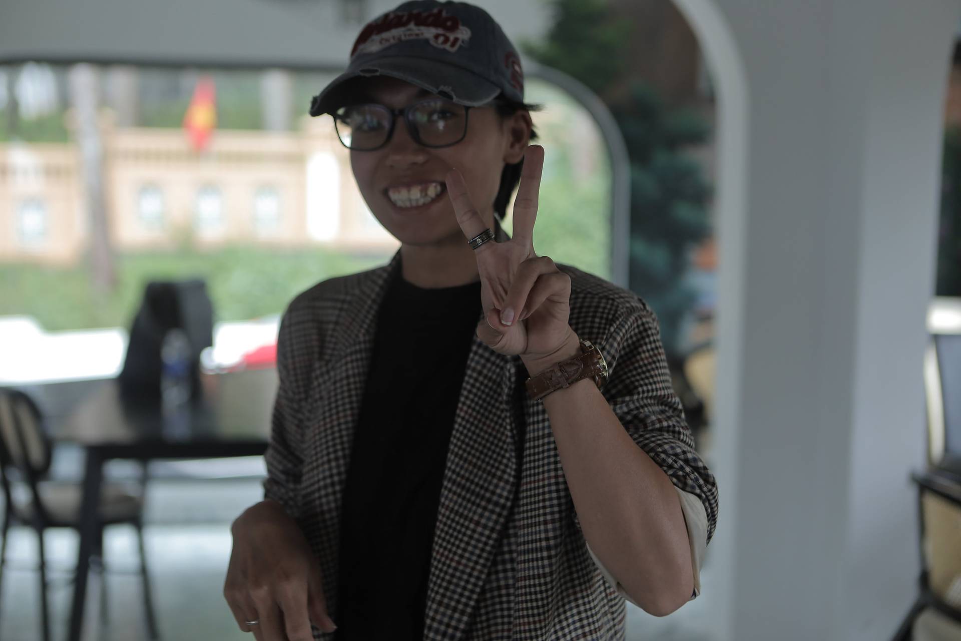 A person in Đà Lạt, Vietnam, making a peace sign with a smile while wearing casual attire and a cap, set against a scenic backdrop of a building and greenery.